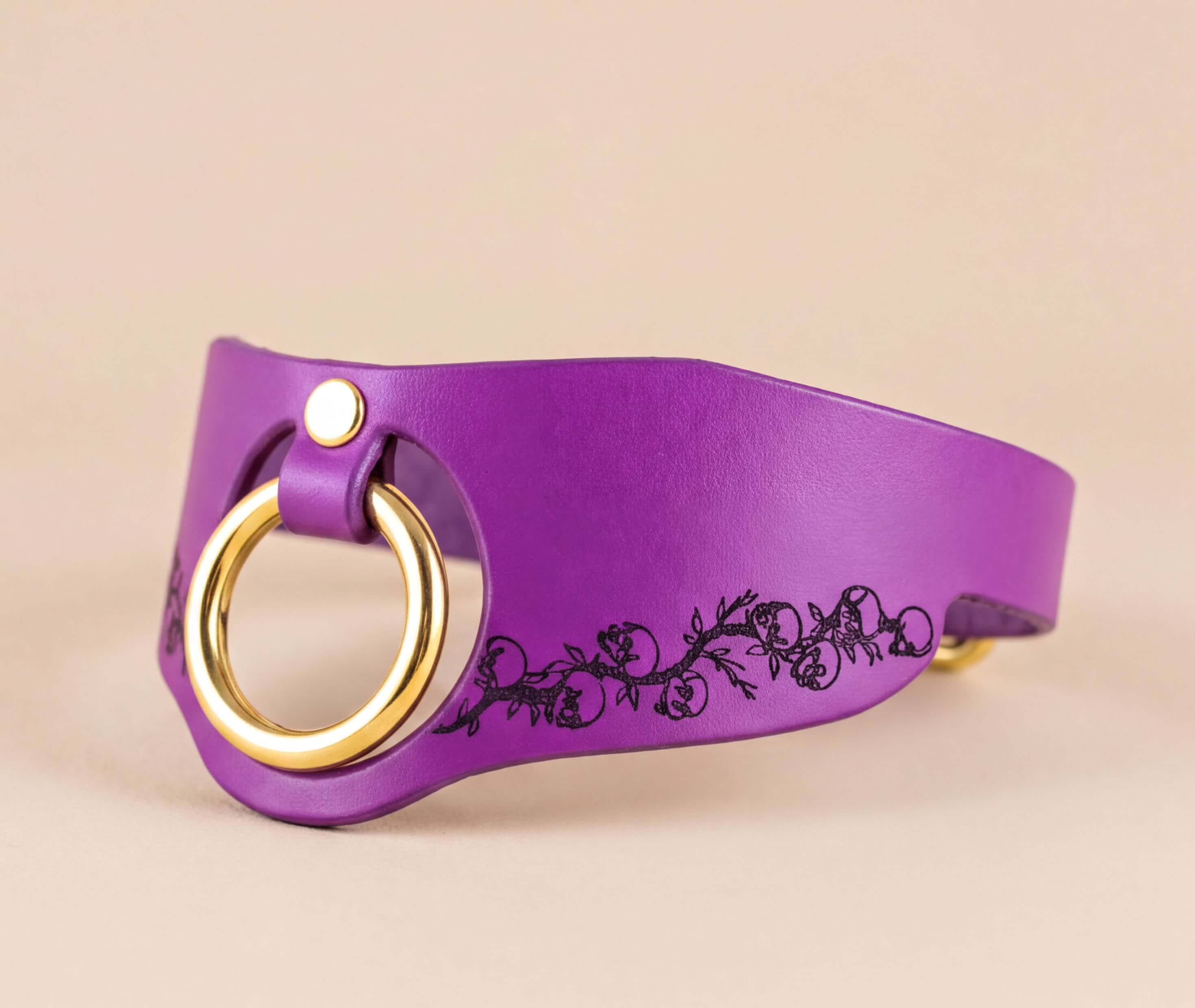 A purple leather choker necklace with a gold-colored ring and a black floral design on the side.