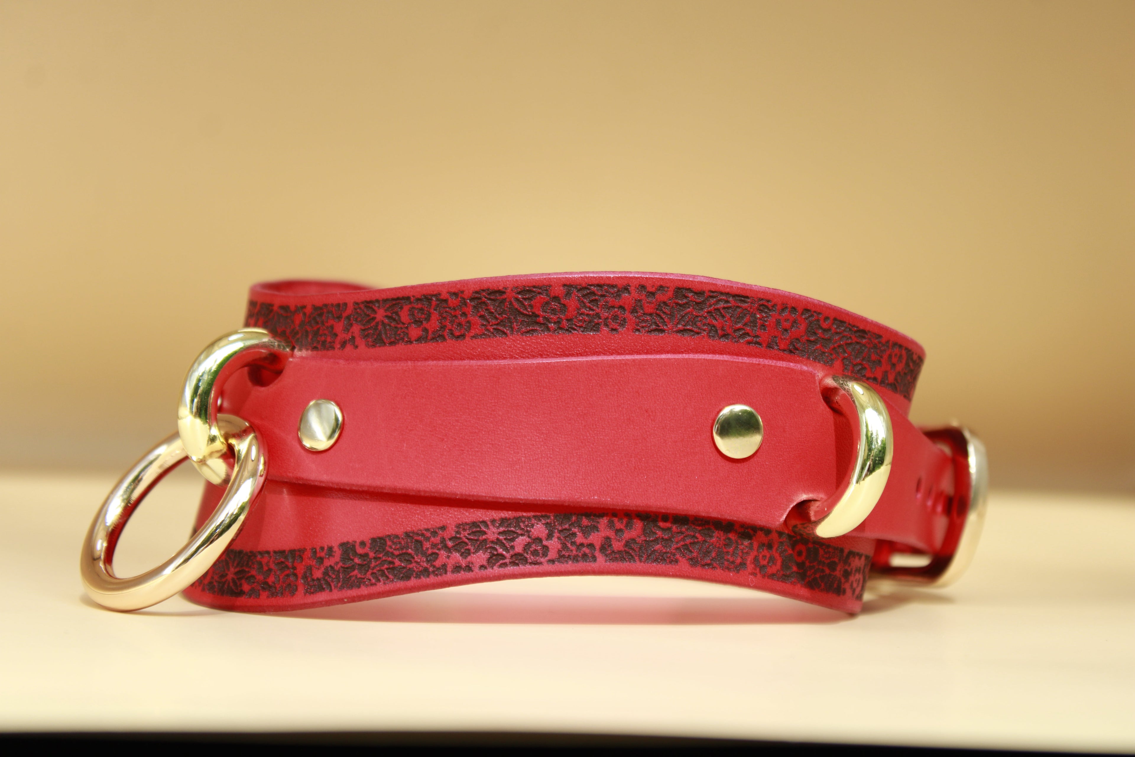 A red leather choker with a floral pattern and two gold-colored buckles is displayed on a light-colored surface.