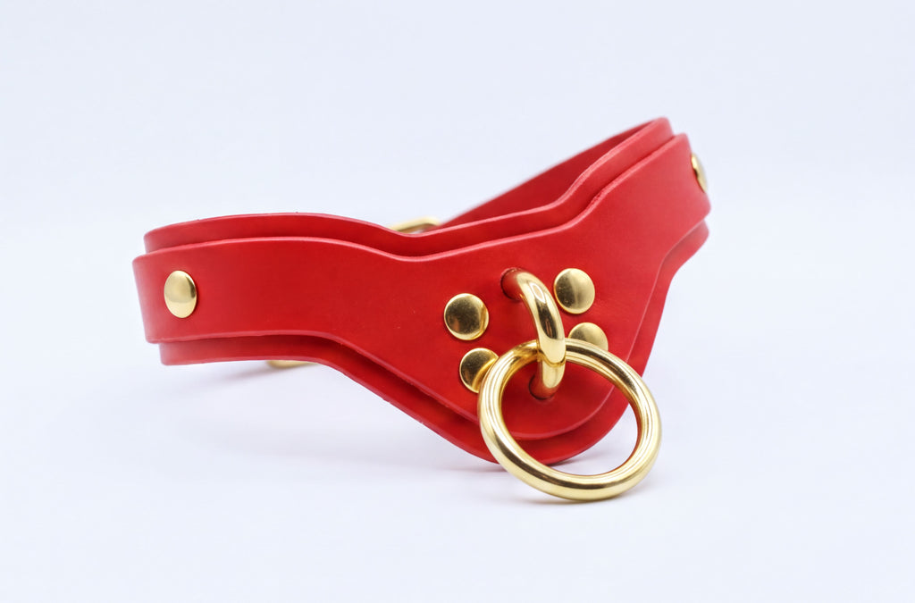 A red leather collar with a gold ring and studs, featuring a curved shape and a metallic appearance.