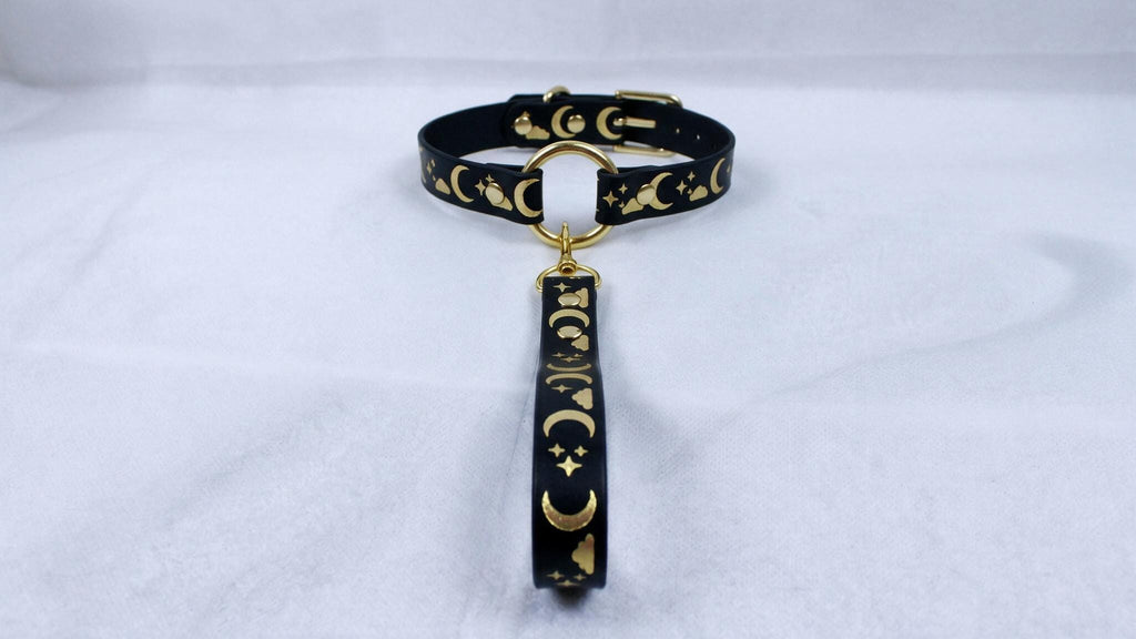 A black collar with gold moon and star designs, featuring a gold-colored buckle and a dangling charm.