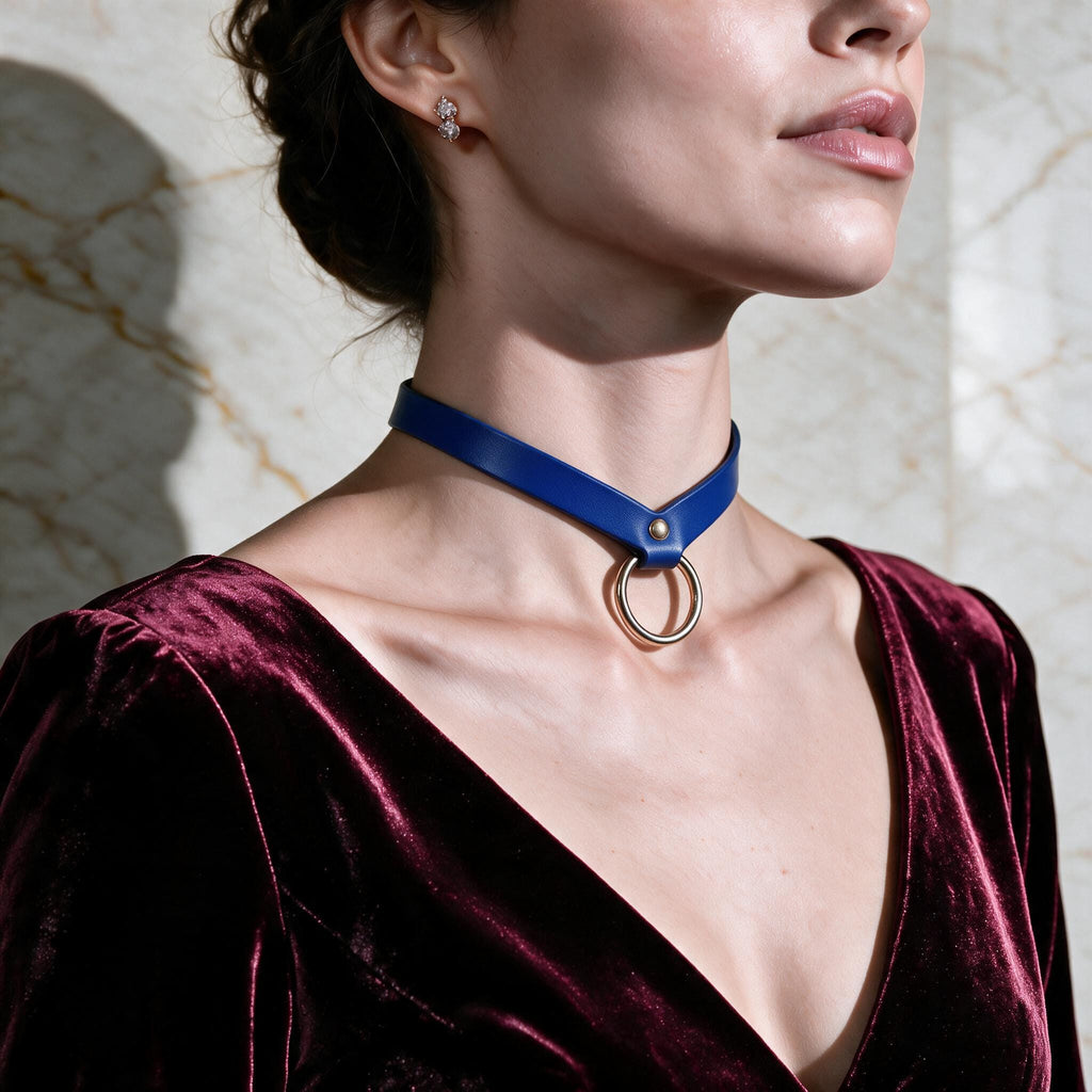 A woman wearing a blue choker necklace with a gold ring, standing against a marble wall.