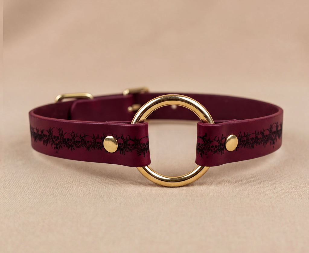 A burgundy leather choker collar with a gold ring and a black floral pattern on the strap.