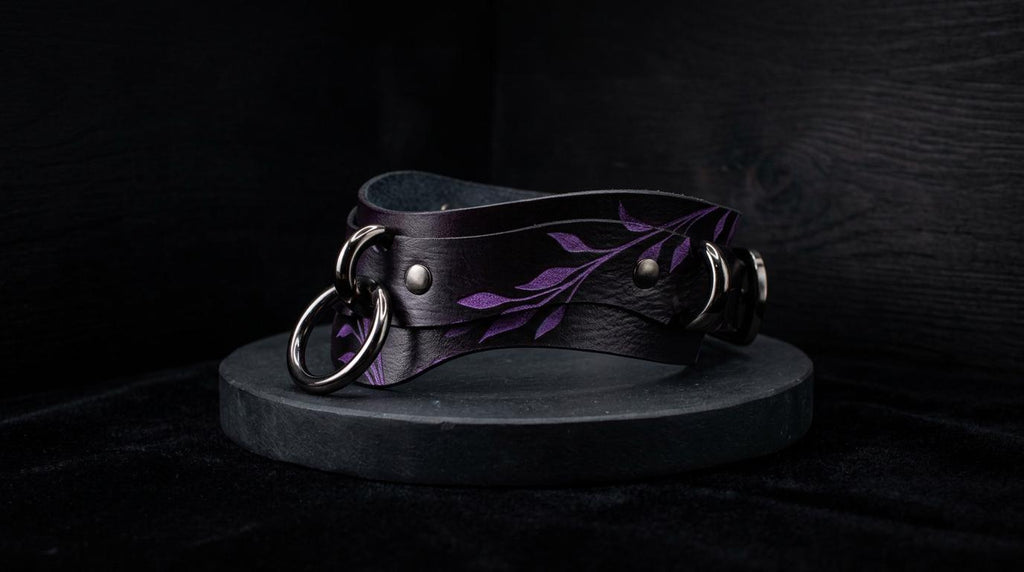 A black leather choker necklace with purple leaf designs and metal rings is displayed against a dark background.