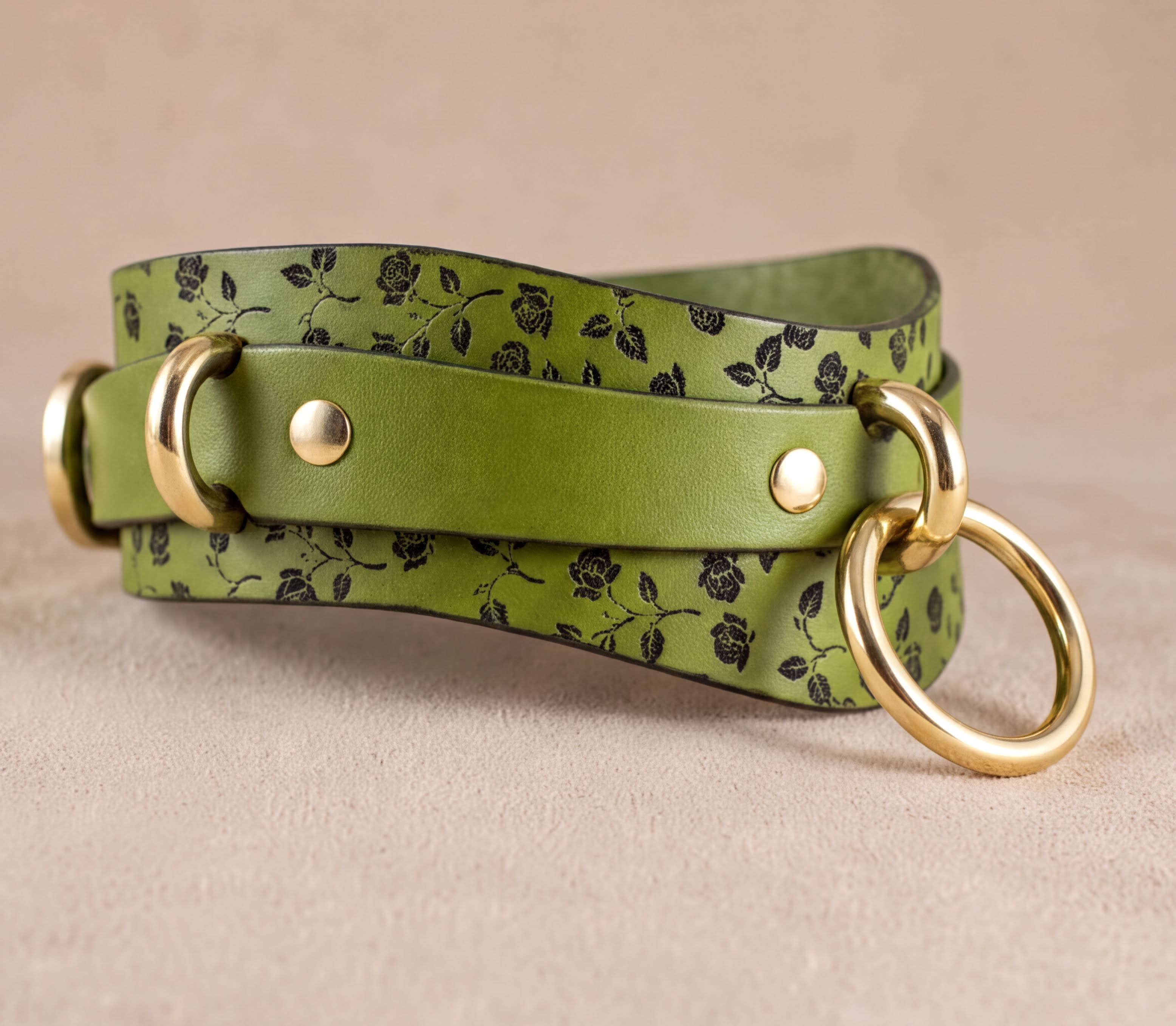 A green leather strap with a floral pattern and two gold rings on it.