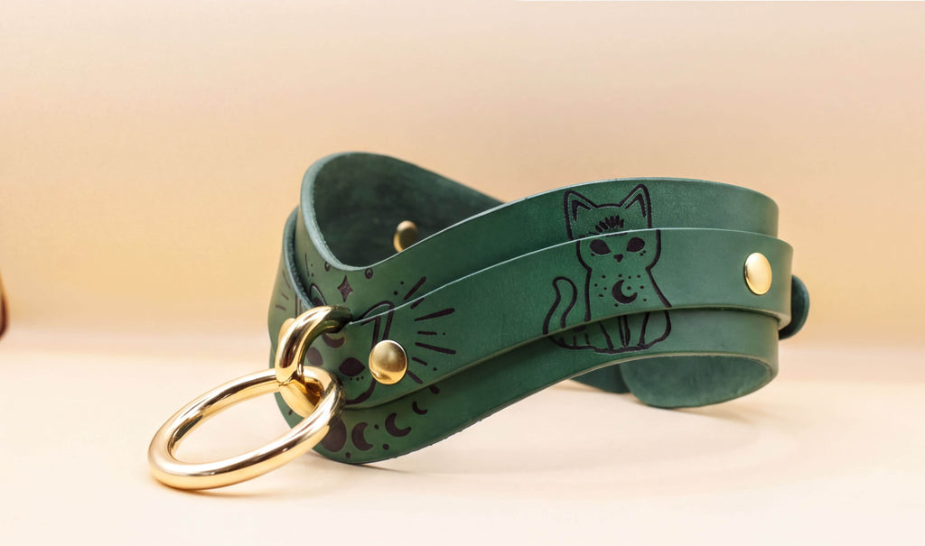 A green leather cat collar with a gold ring and a cat illustration on it.