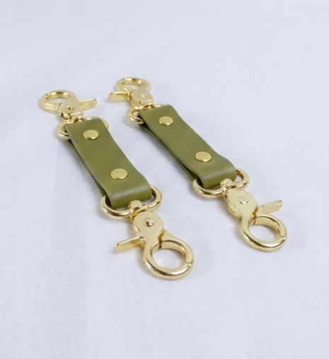 A pair of gold-colored leather straps with brass-colored metal hooks attached to them, placed on a white surface.