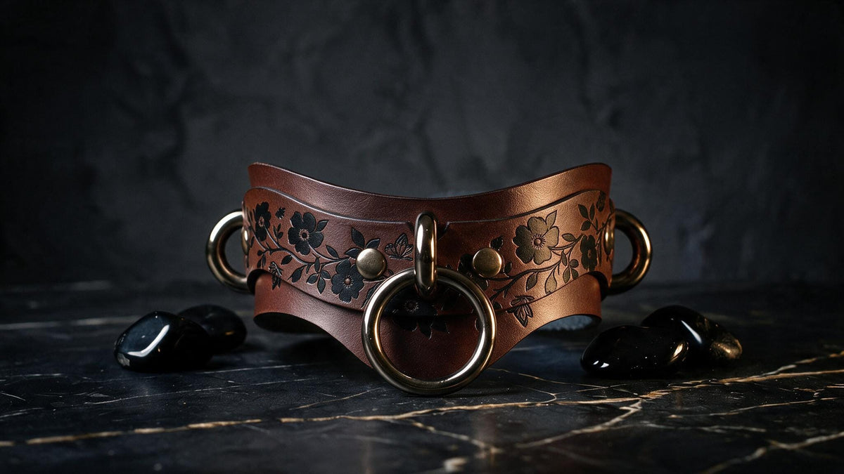 A brown leather choker necklace with floral designs and metal rings is displayed on a dark surface, accompanied by black stones.