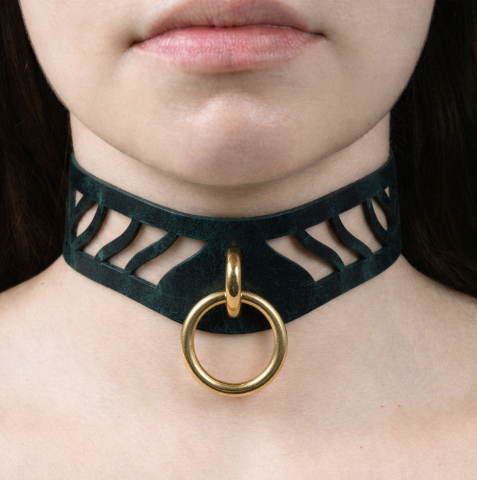 A close-up of a woman's neck and chin area, featuring a black choker necklace with a gold ring and a cutout design.