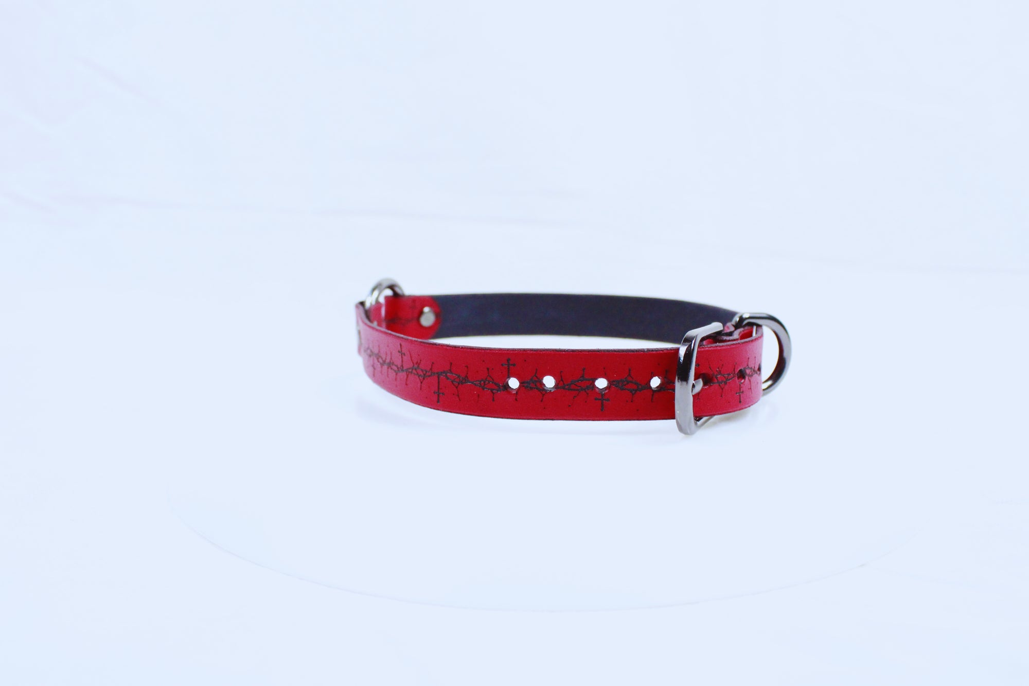 A red leather dog collar with a black buckle and a patterned design on the strap is shown against a white background.