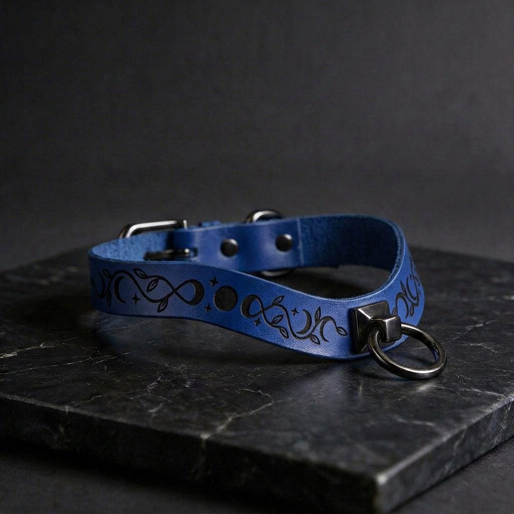 A blue leather collar with a metal ring and a decorative pattern on the strap is displayed on a black marble surface.