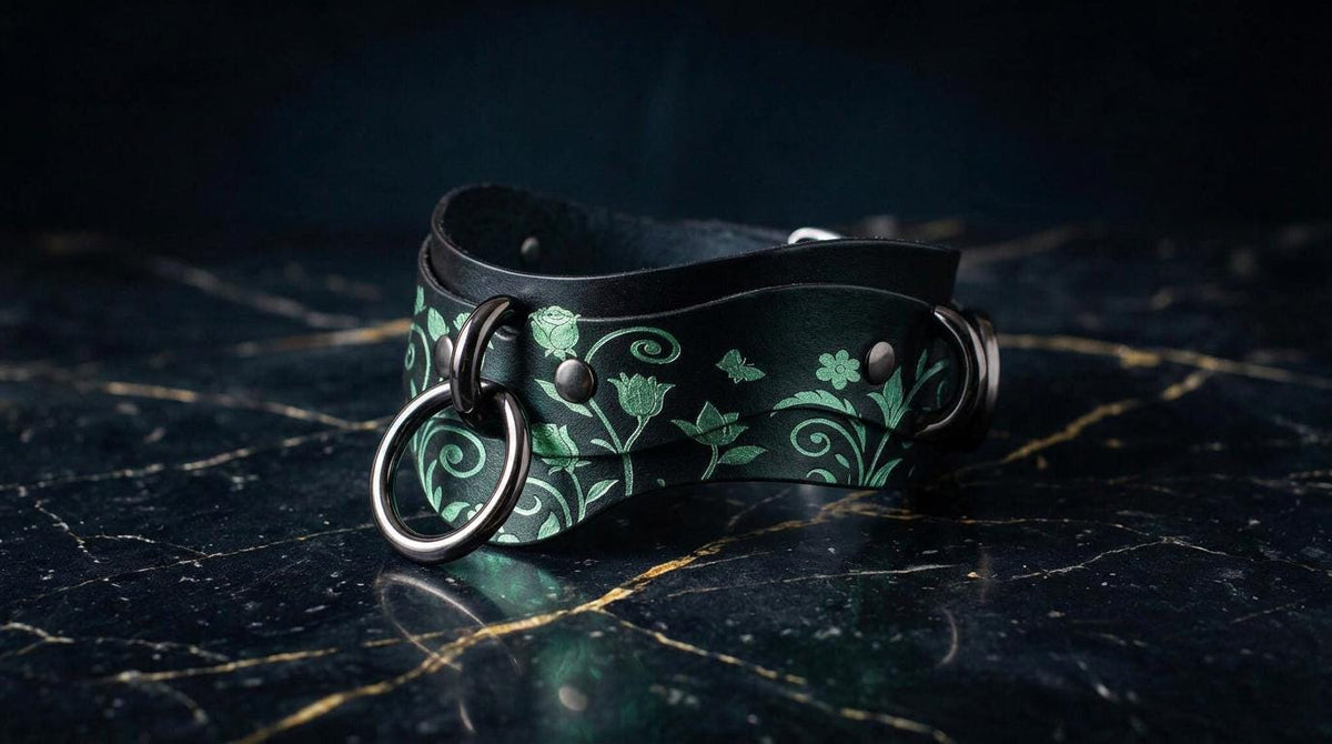 A black leather choker necklace with a green floral pattern and metal rings on the front, resting on a dark marble surface.