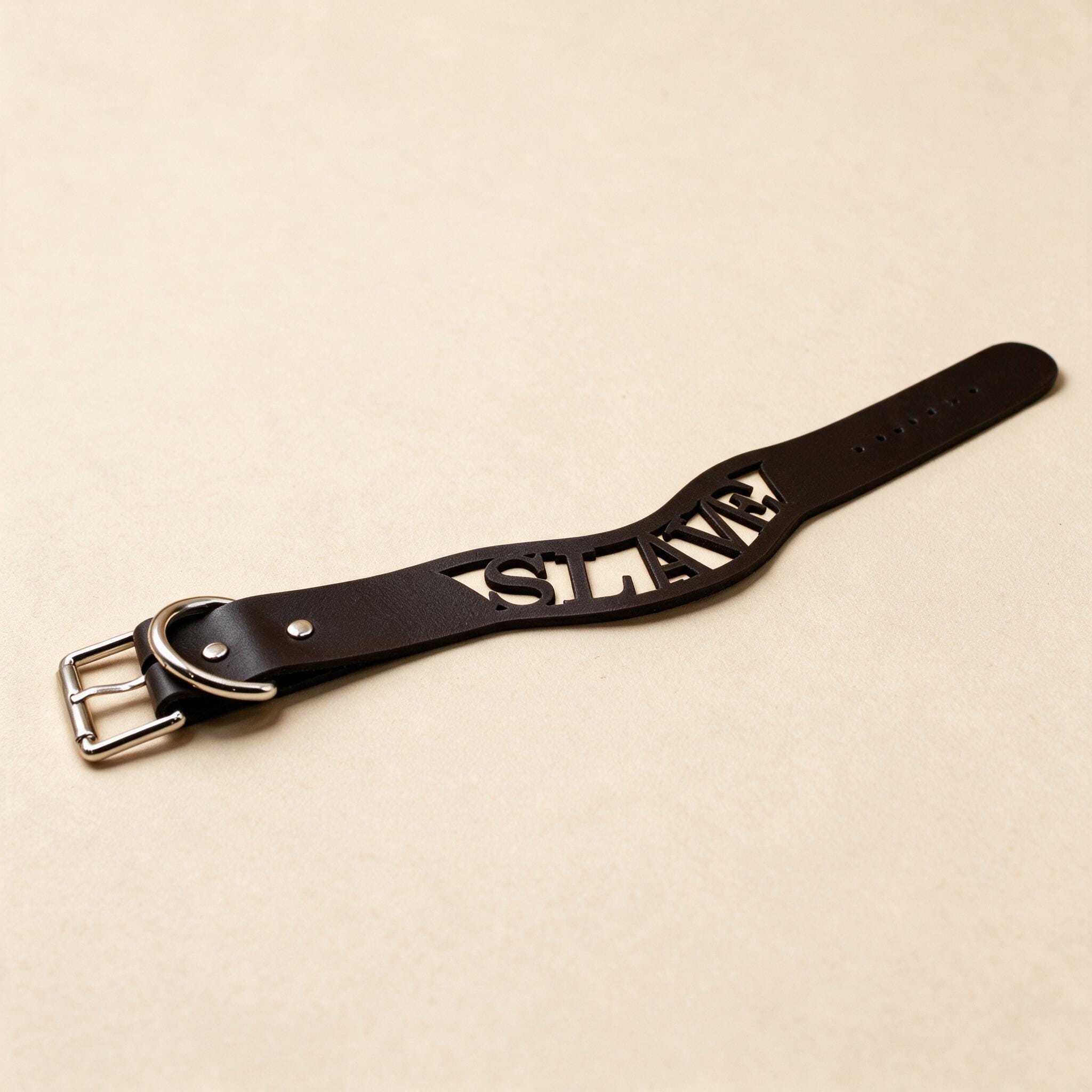 A black leather strap with the word "SLAVE" cut out in the center, lying on a beige surface.