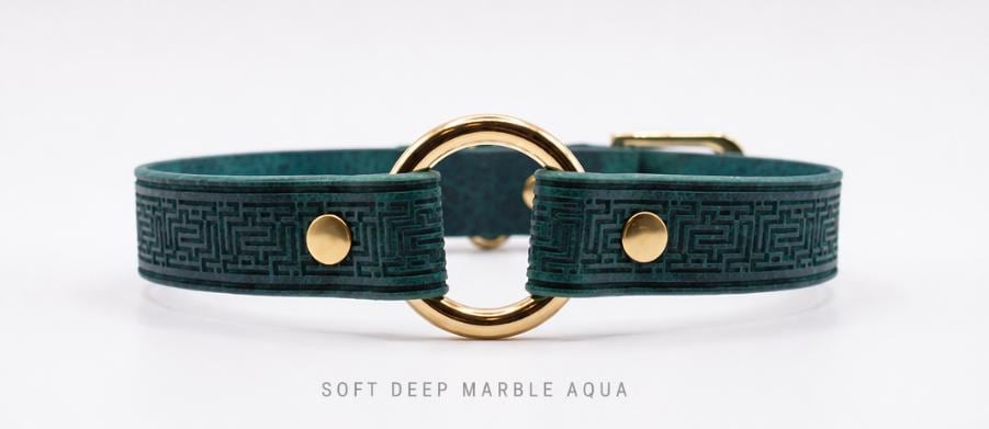 A close-up image of a green leather collar with a gold-colored metal ring and two gold-colored metal buttons.