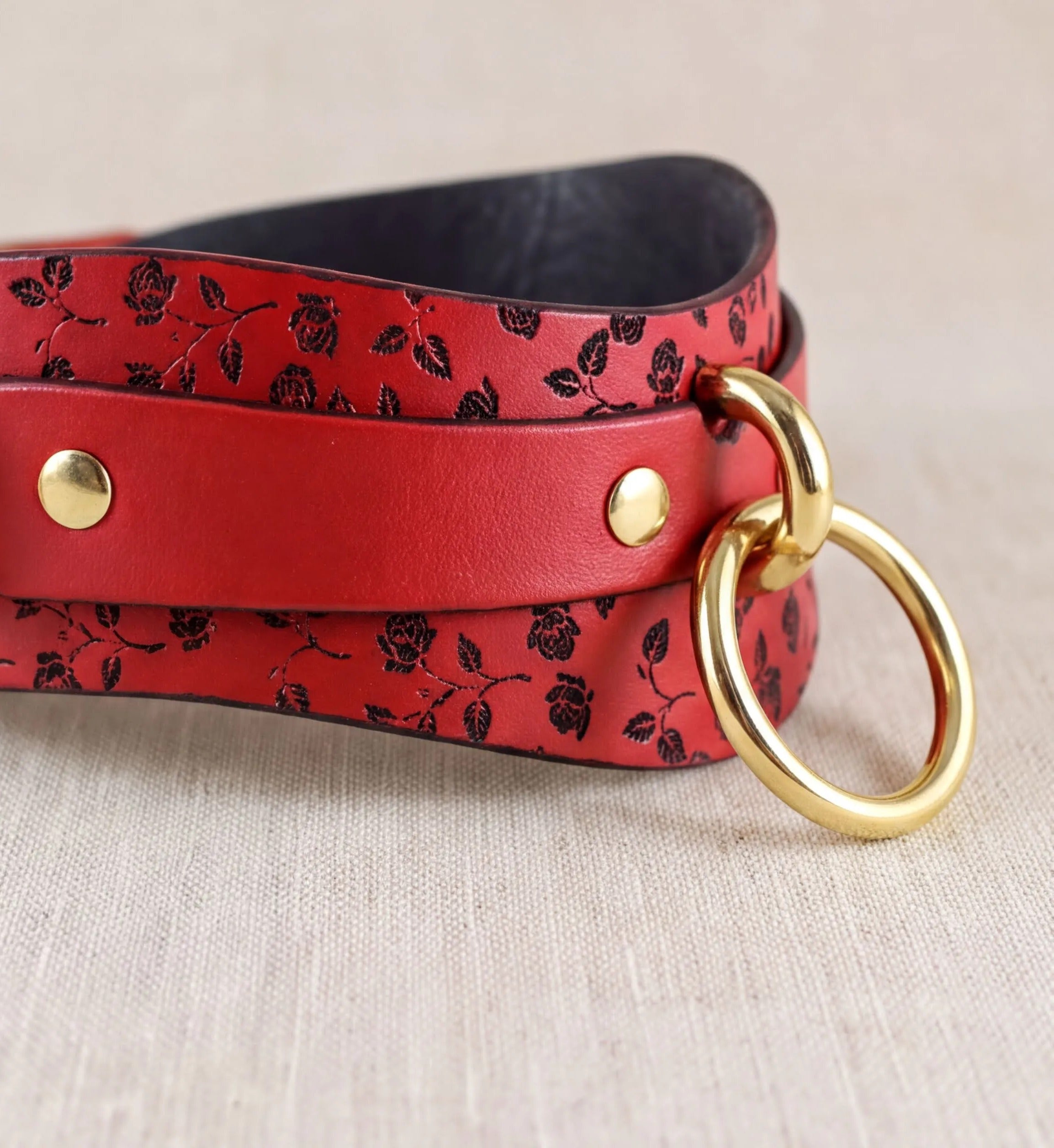 A red leather strap with a floral pattern and two gold rings on the ends, placed on a beige surface.