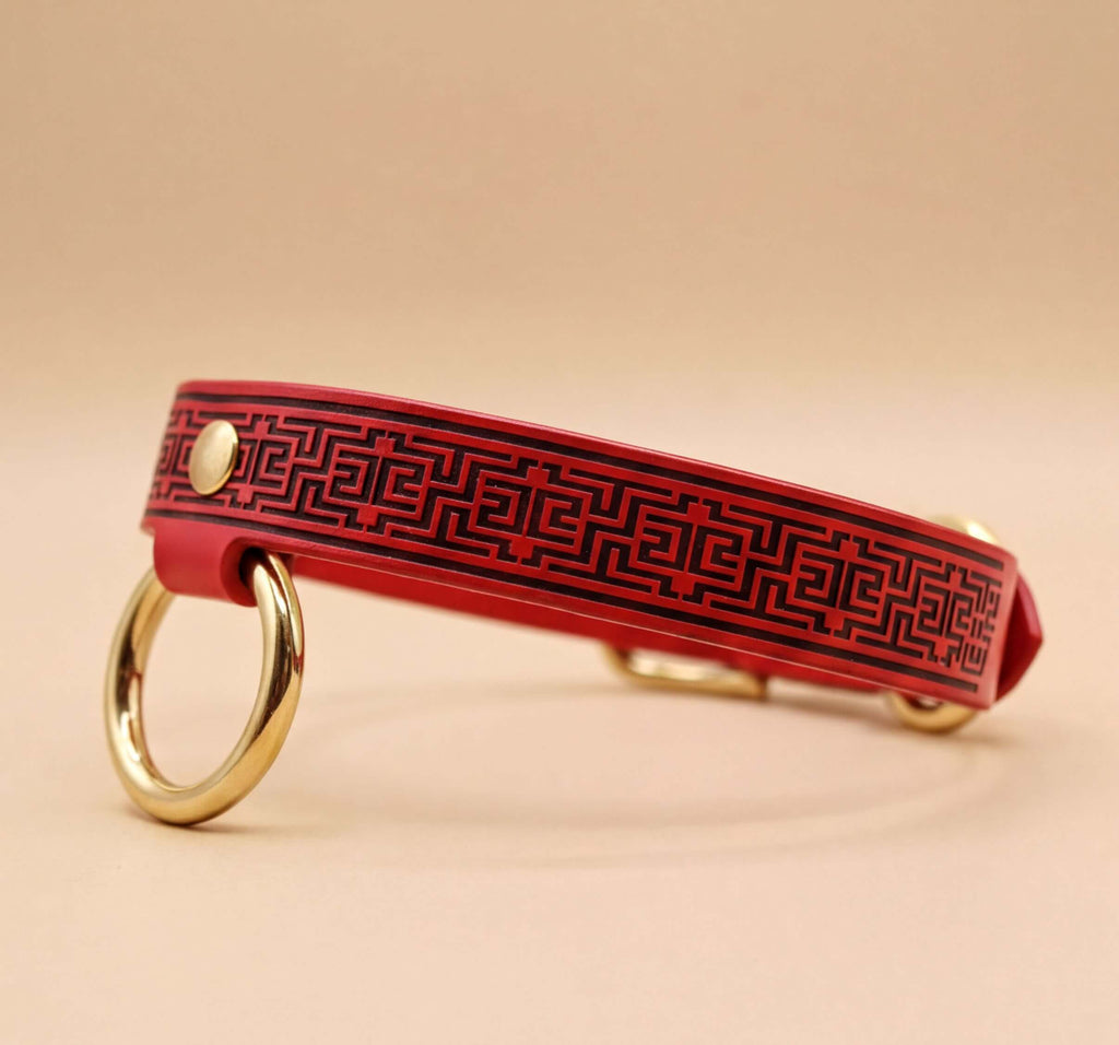 A red leather collar with a black geometric pattern and two gold-colored rings on the sides.