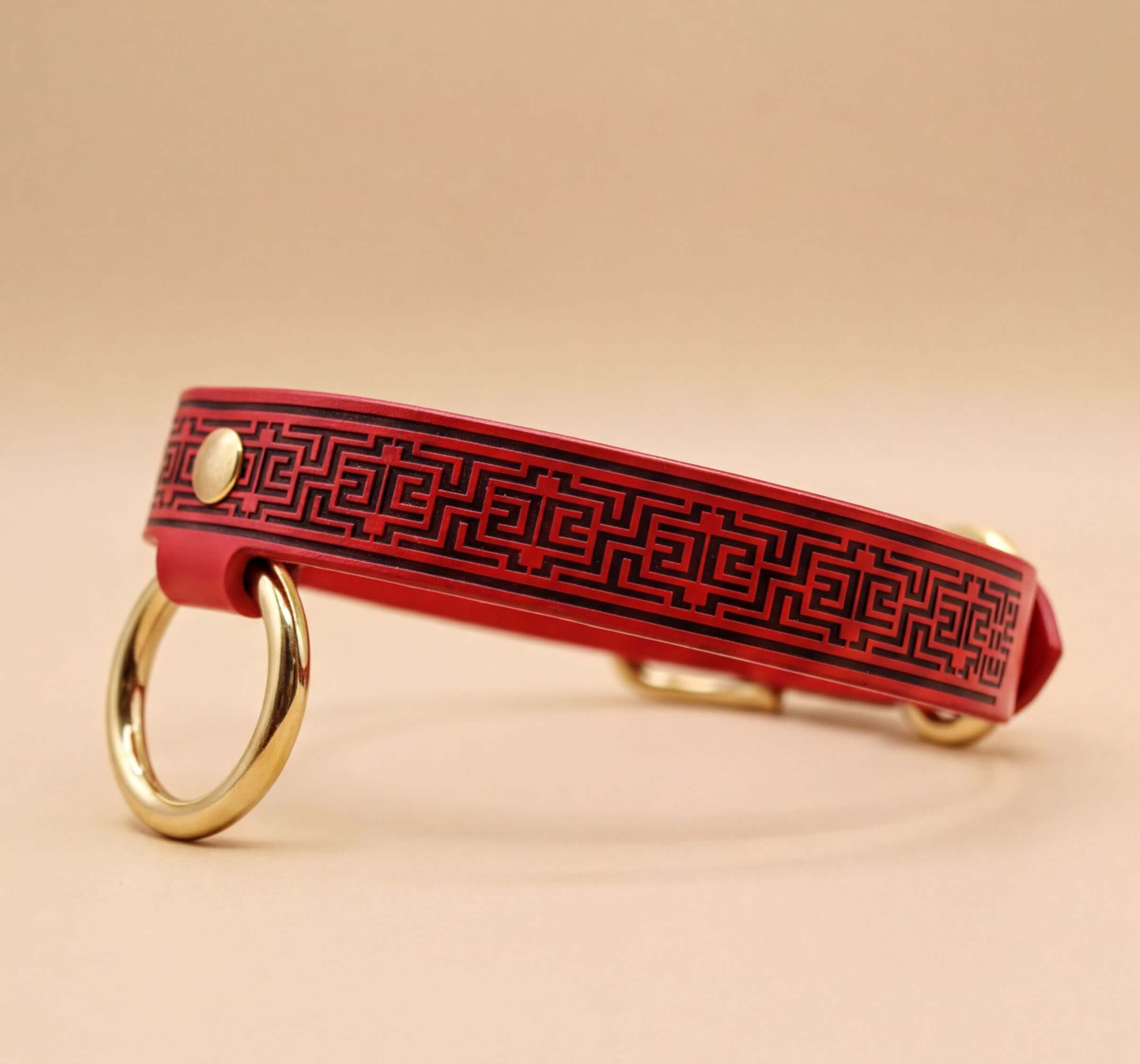 A red leather collar with a black geometric pattern and two gold-colored rings on the sides.