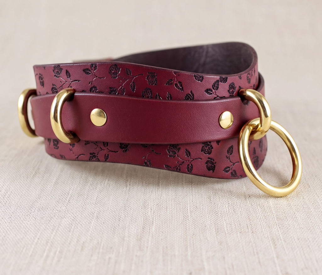 A burgundy leather strap with a floral pattern and two gold-colored metal rings on the ends, resting on a beige background.