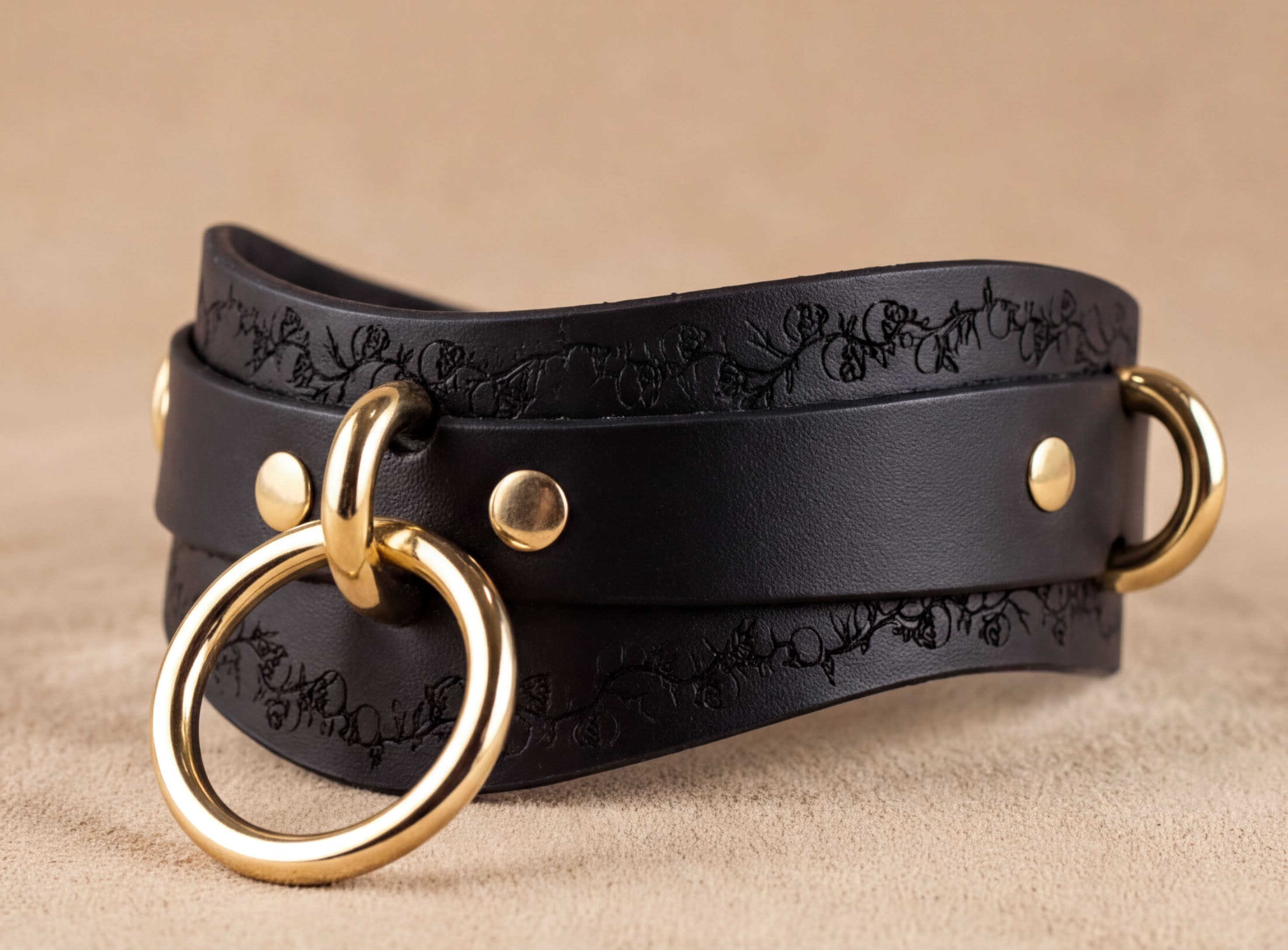 A black leather choker necklace with gold metal rings and a decorative floral pattern on the leather.