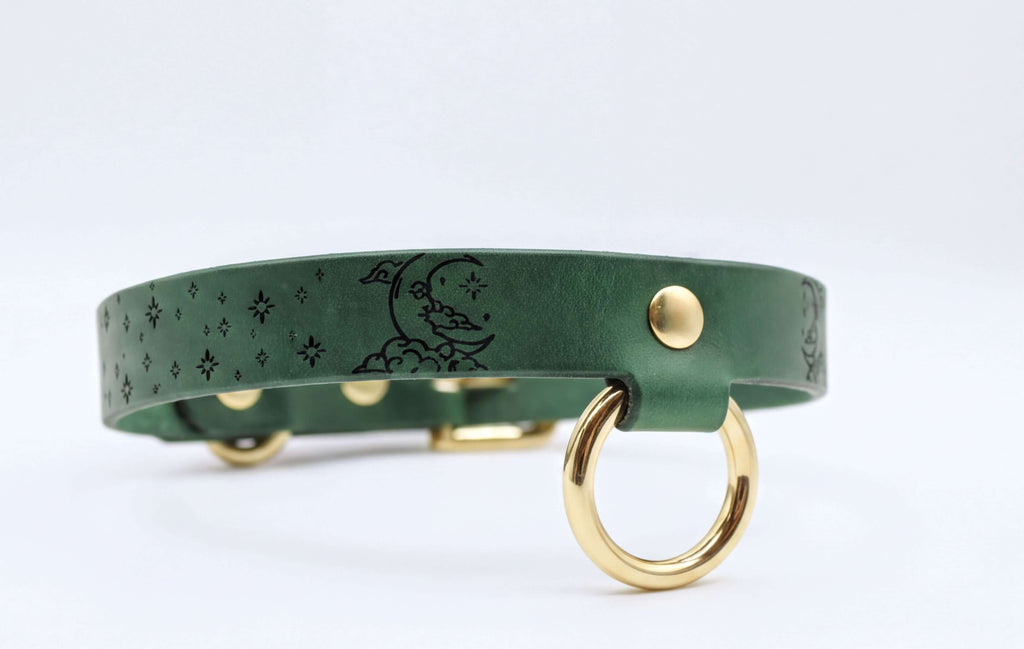 A green leather collar with a moon and star design, featuring a gold-colored metal ring on the front.