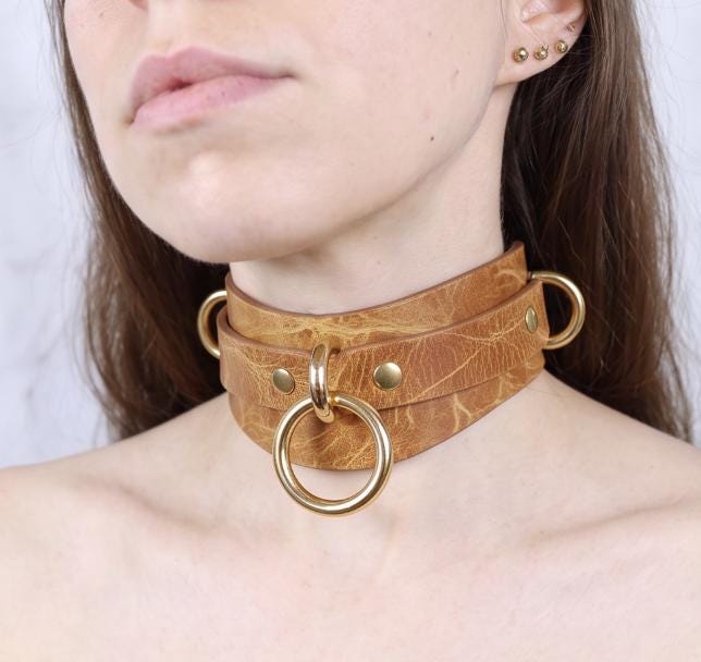 A close-up of a woman's neck and chest area wearing a brown leather choker necklace with gold-colored rings.