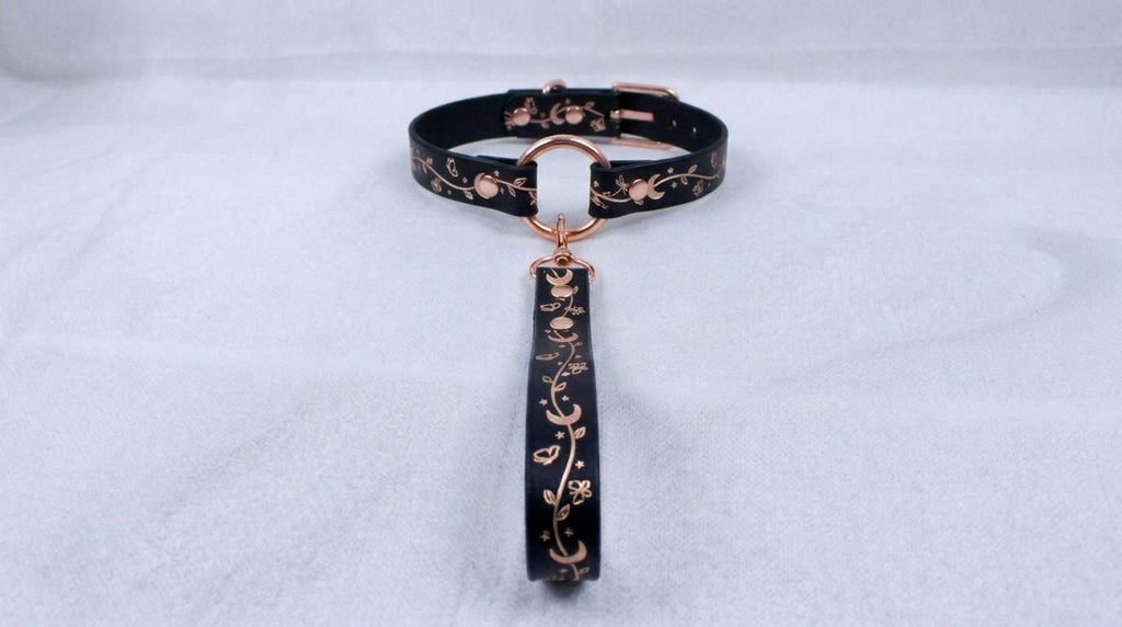 A black collar with a gold-colored metal clasp and a decorative handle attached to it, resting on a white surface.