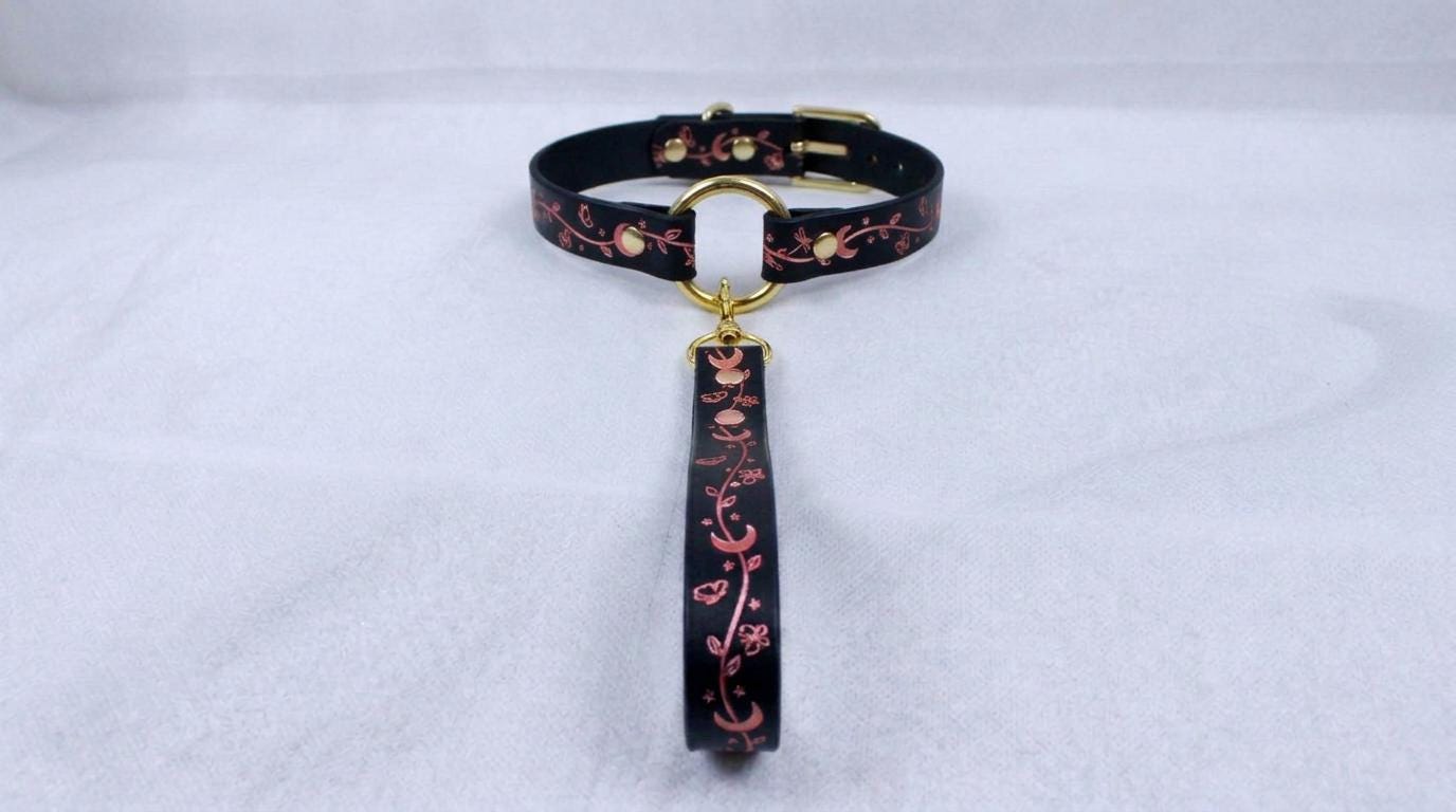 A black collar with a gold-colored buckle and a floral pattern on the strap is displayed against a white background.
