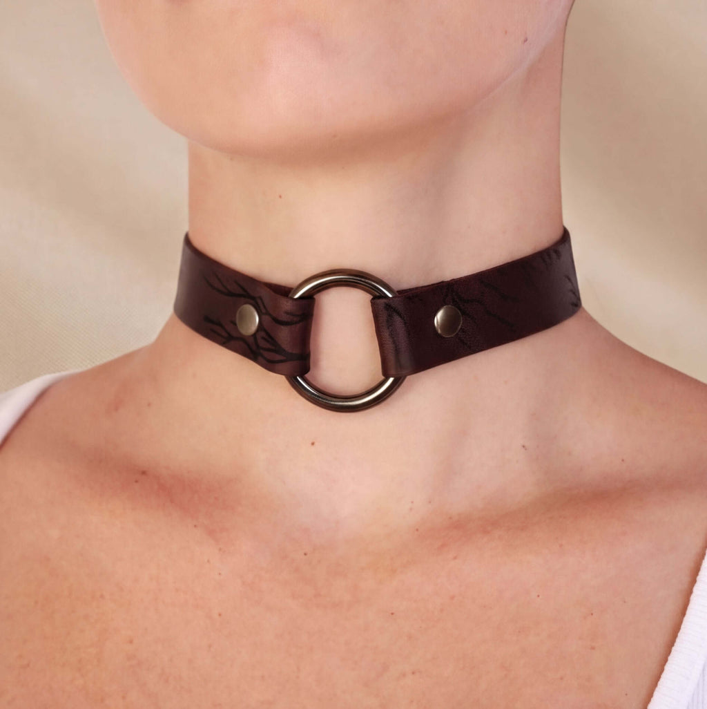 A close-up of a person's neck wearing a dark brown leather choker necklace with a circular metal ring.