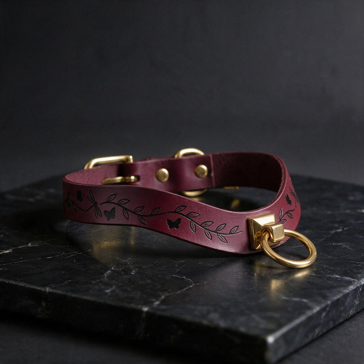 A burgundy leather collar with a leaf and butterfly design, featuring a gold-colored metal ring and buckle, is displayed on a black marble surface.