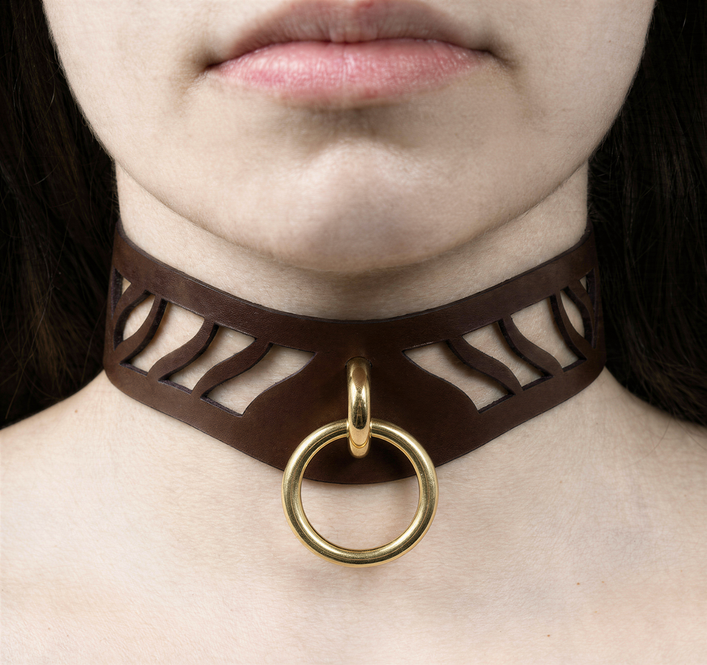 A close-up of a person's neck and chest area, featuring a brown leather choker necklace with a gold ring and a cutout design.