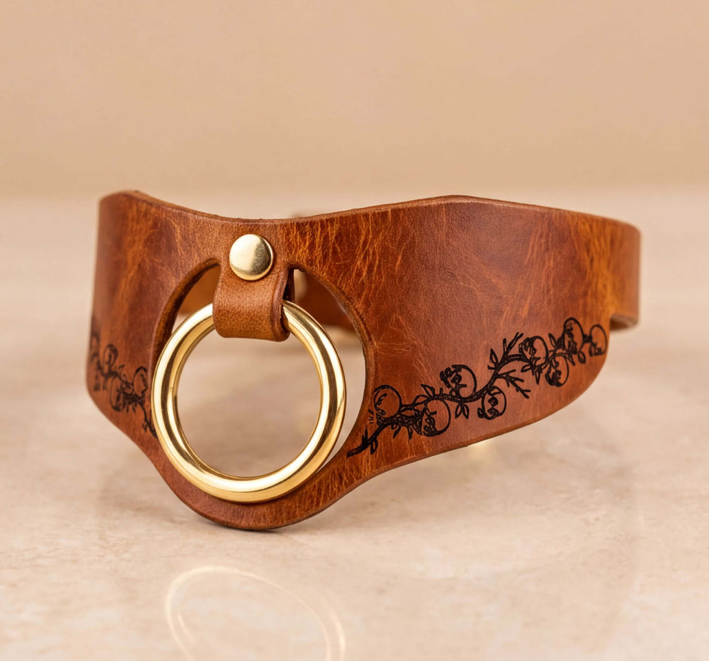 A brown leather choker necklace with a gold-colored ring and a floral design on the side.