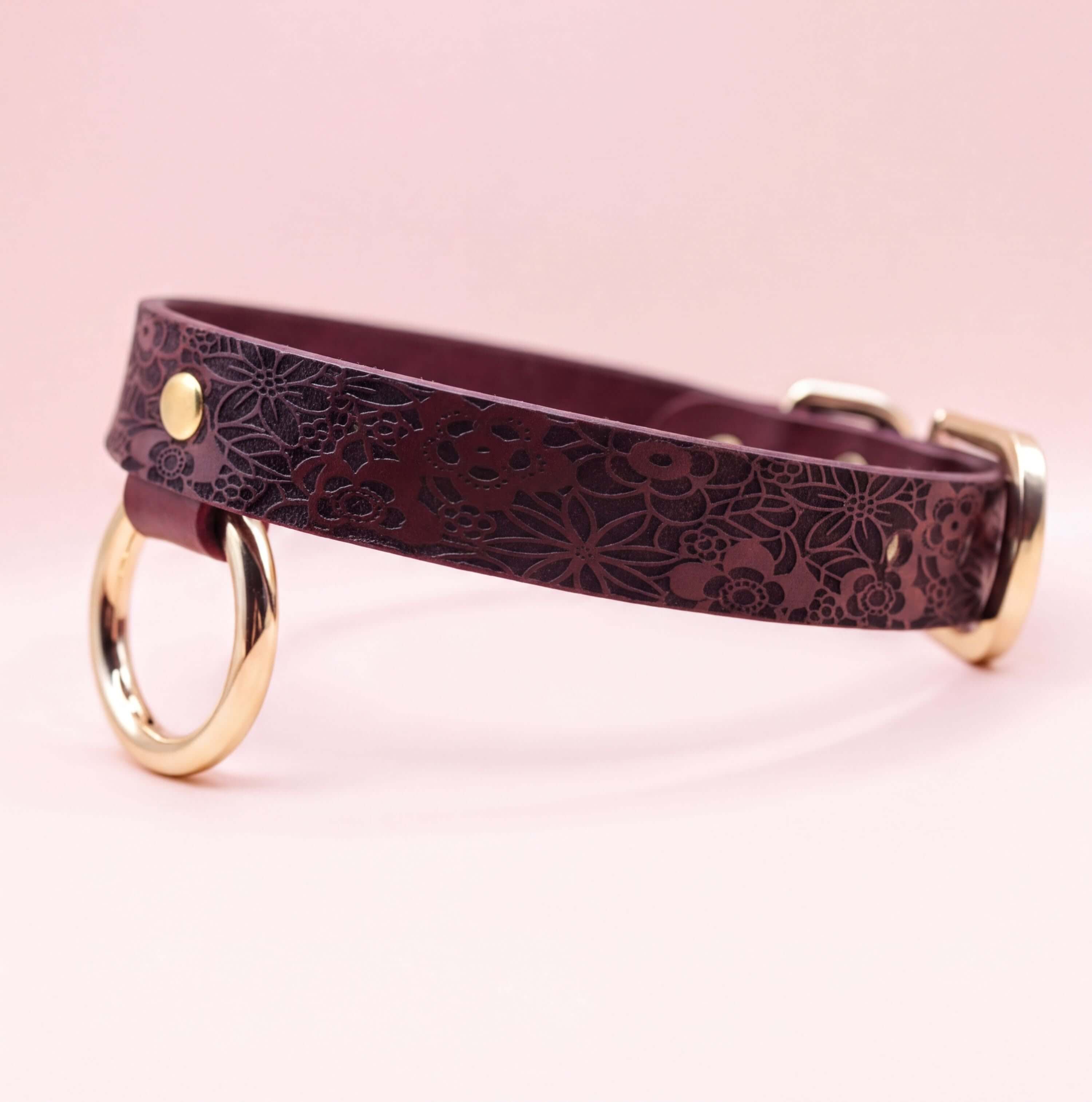 A burgundy leather collar with a floral pattern and two gold-colored rings on the front.