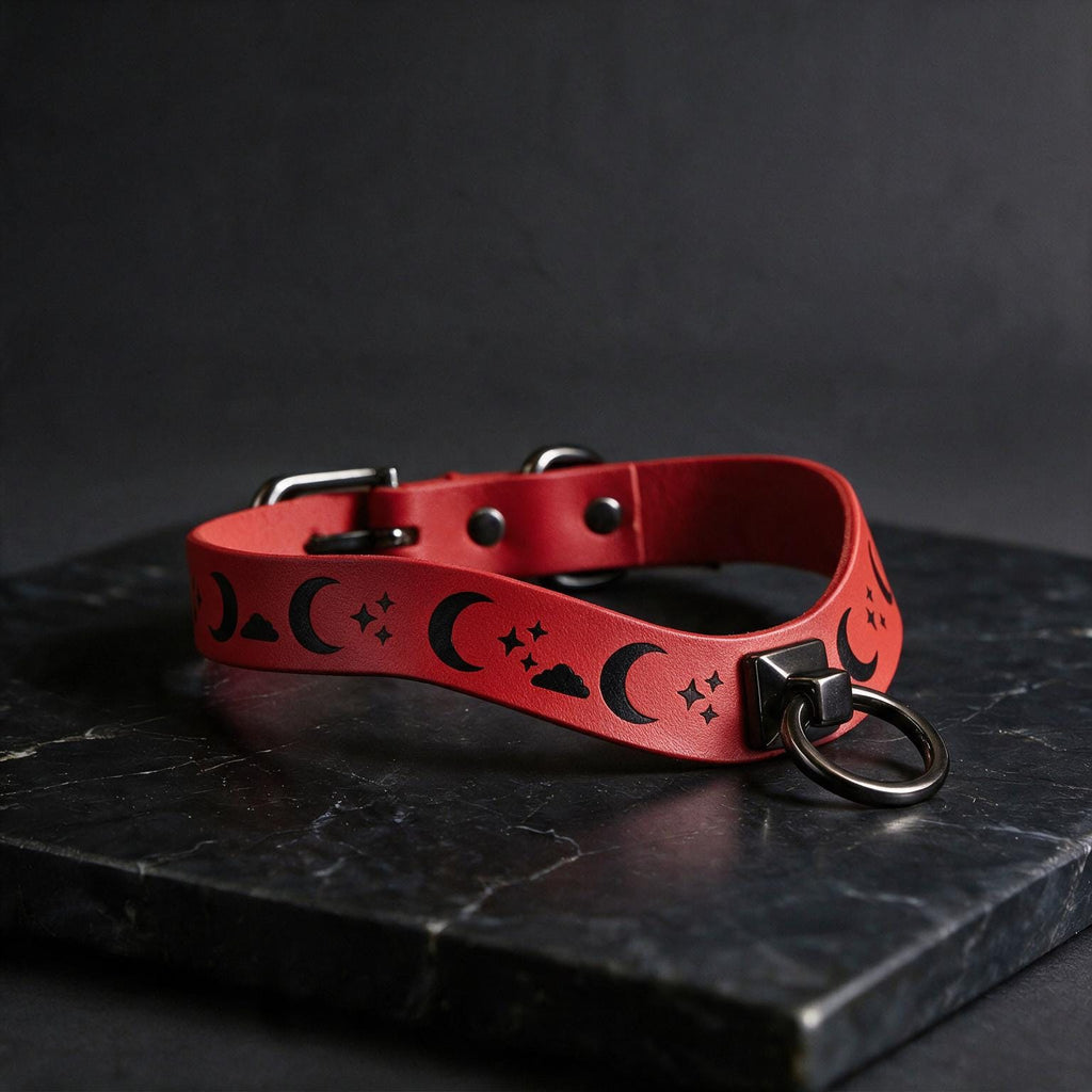 A red leather collar with a moon and star pattern design, featuring a metal ring and buckle, is displayed on a black marble surface.