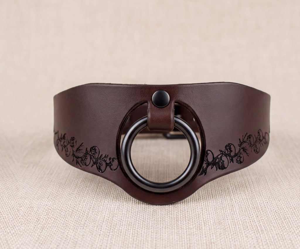 A brown leather collar with a black circular ring and a decorative pattern on the edge.