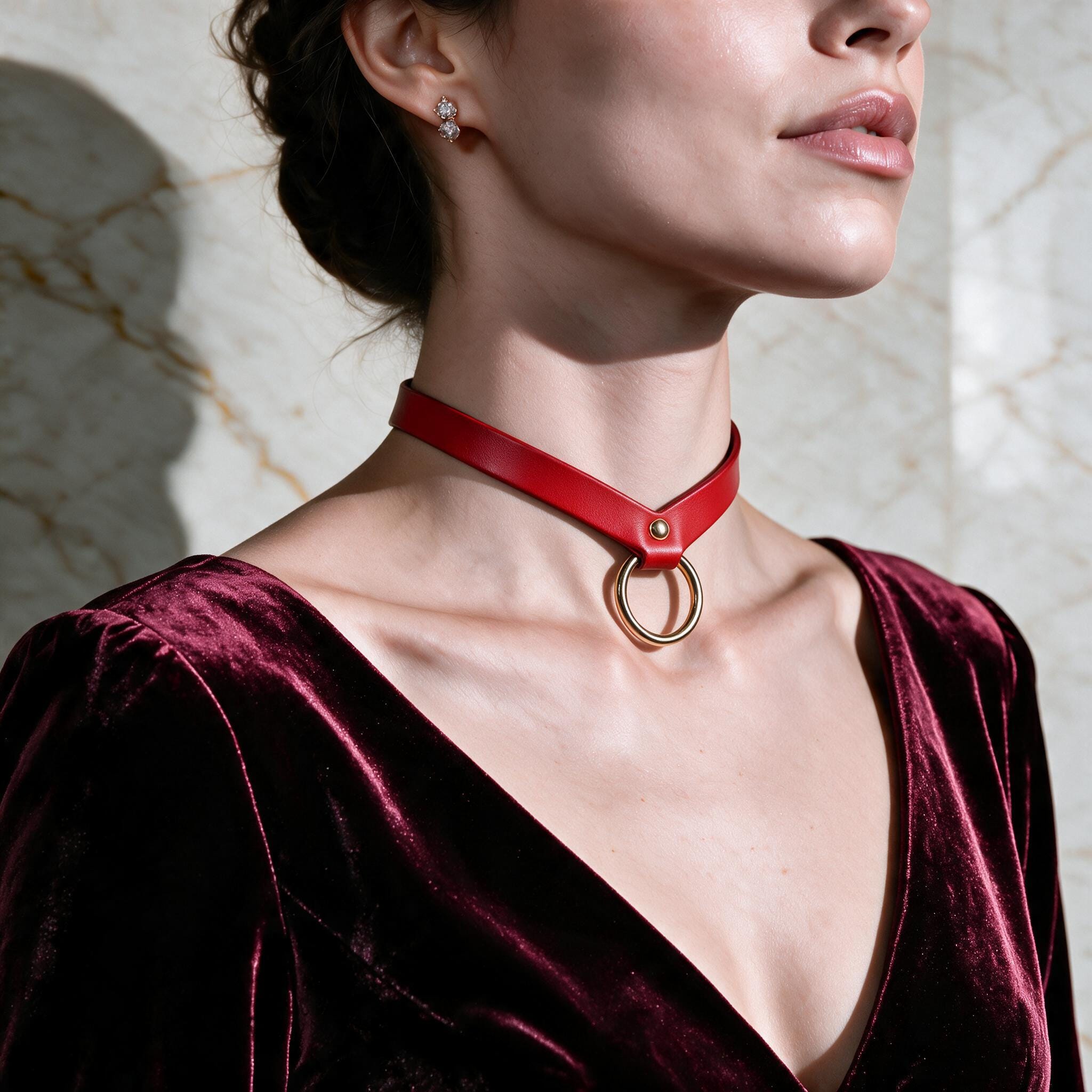 A woman wearing a red velvet dress with a red choker necklace and earrings stands against a marble wall.