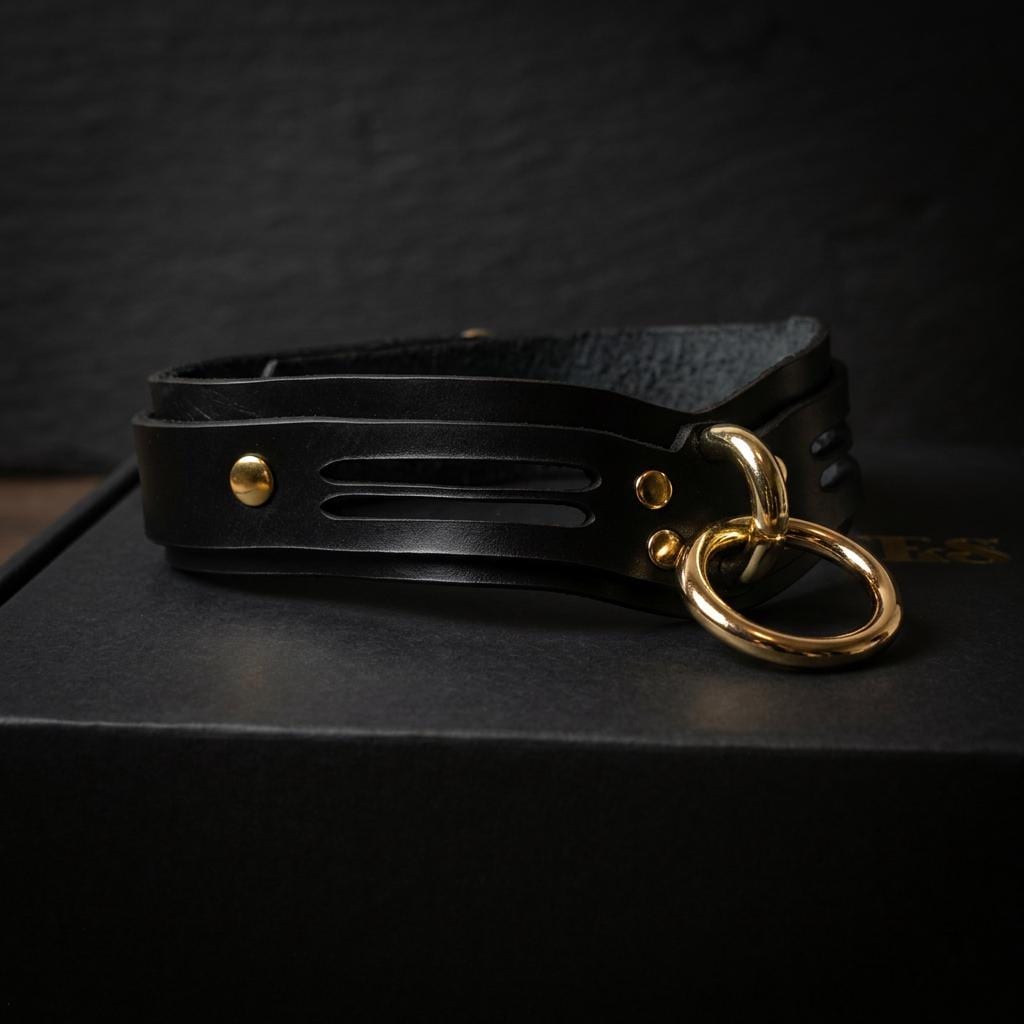 A black leather choker necklace with a gold ring and buckle is displayed on a black surface.