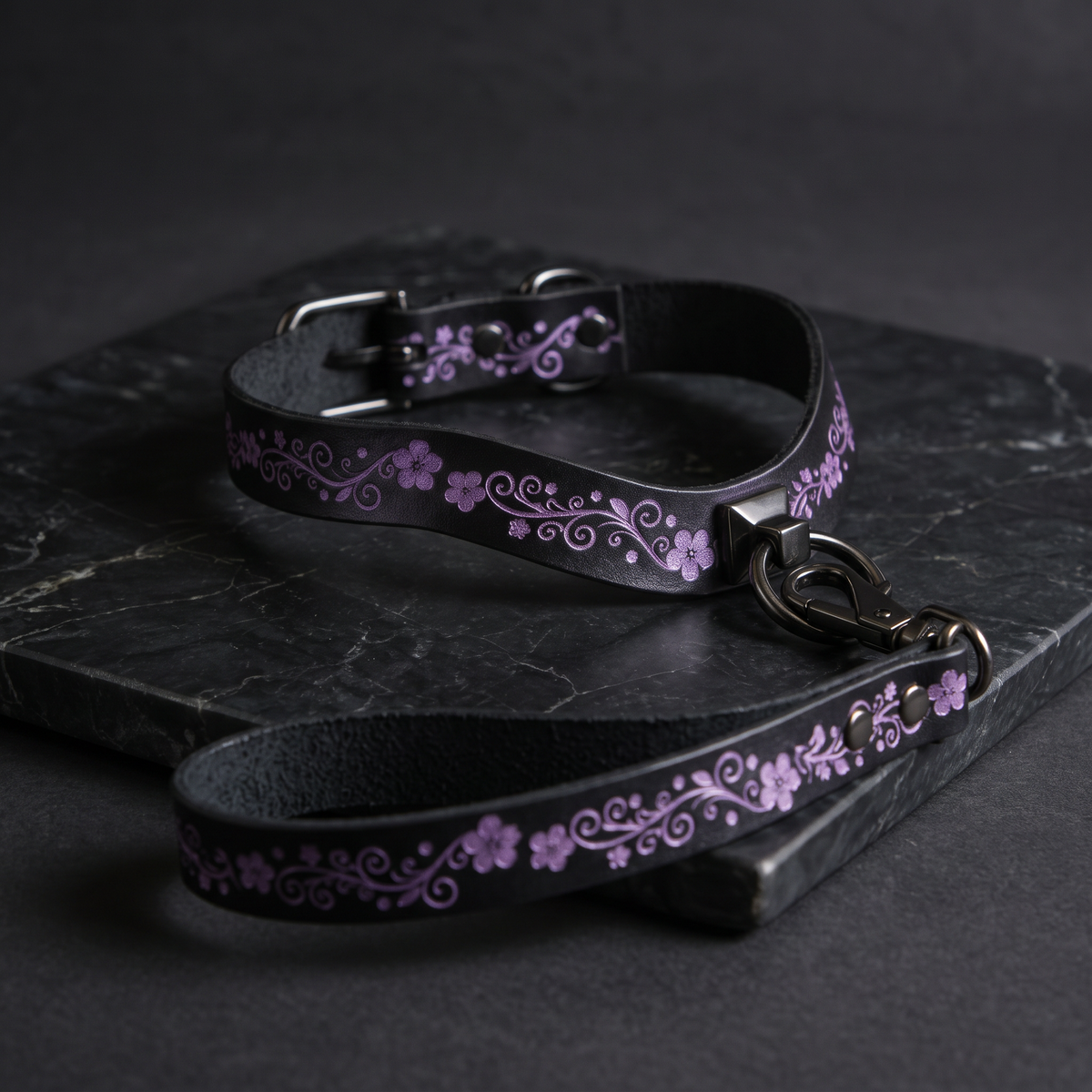 A black leather collar with a floral pattern and a matching leash are displayed on a dark surface.