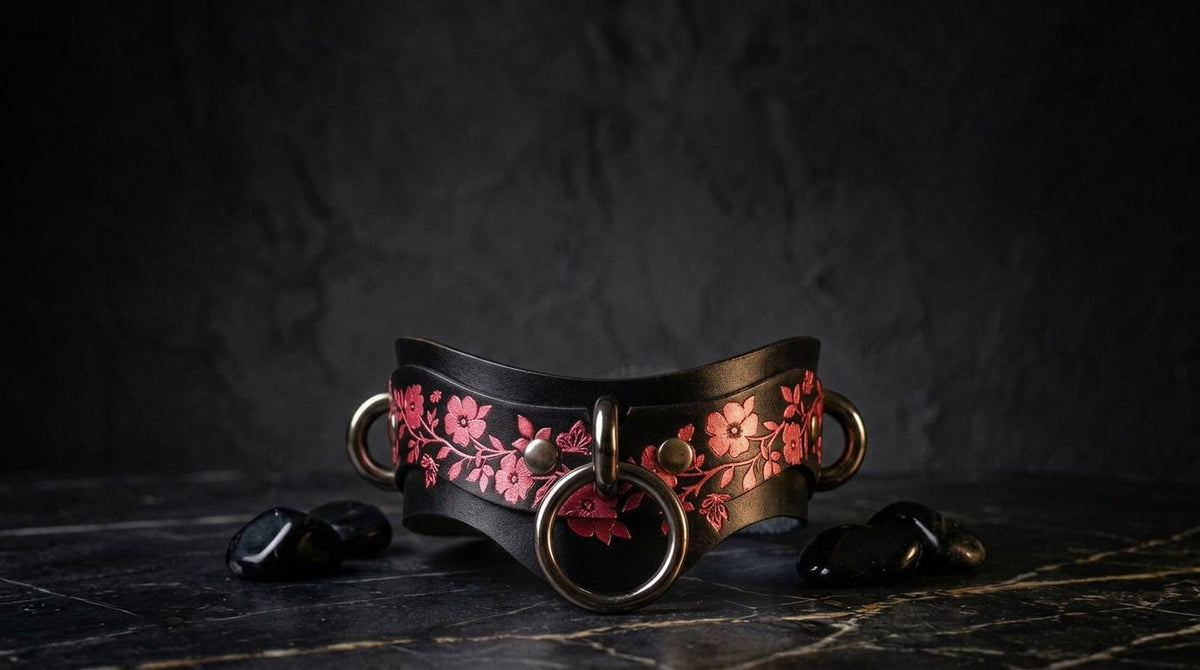 A black leather choker necklace with a floral pattern and metal rings is displayed on a dark surface, accompanied by small black stones.