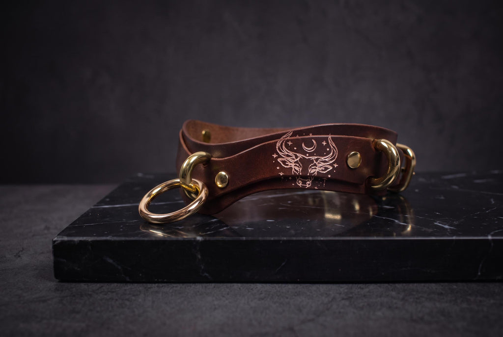 A brown leather choker necklace with gold-colored metal rings and a design on the front is displayed on a black marble surface.