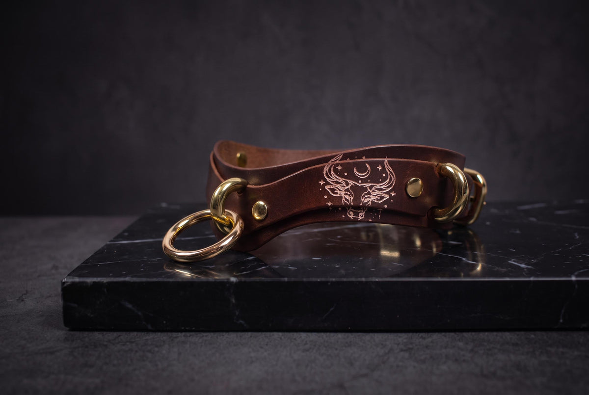 A brown leather choker necklace with gold-colored metal rings and a design on the front is displayed on a black marble surface.