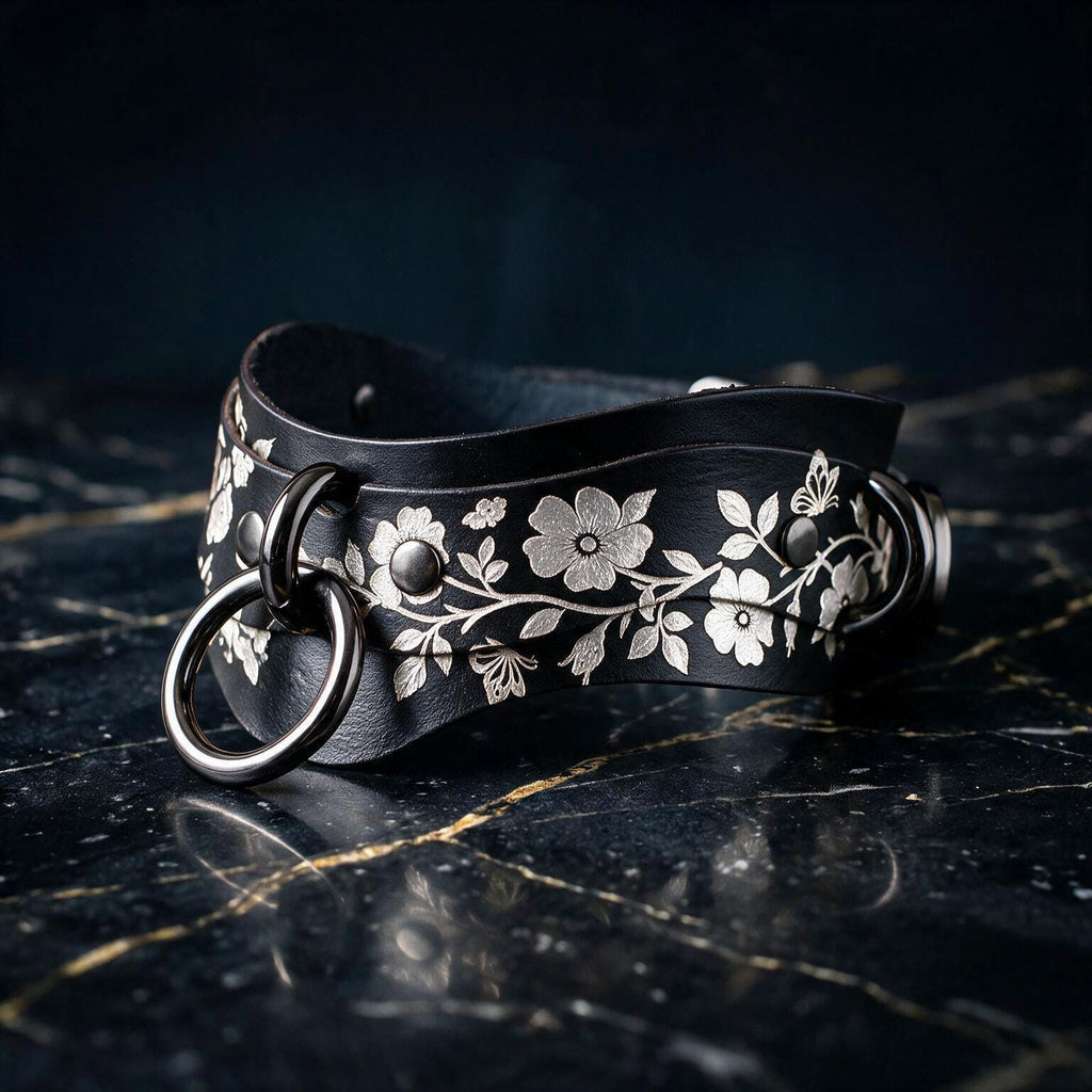A black leather collar with a floral pattern and metal ring on it, resting on a dark surface.