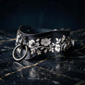 A black leather collar with a floral pattern and metal ring on it, resting on a dark surface.