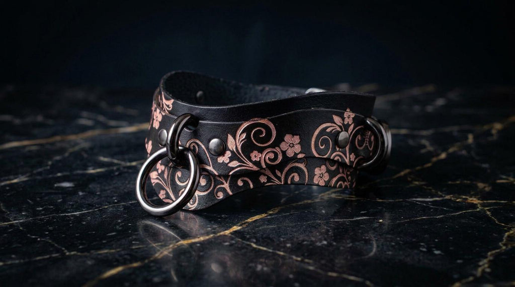 A black leather choker necklace with a floral pattern and metal rings on the ends, resting on a dark marble surface.