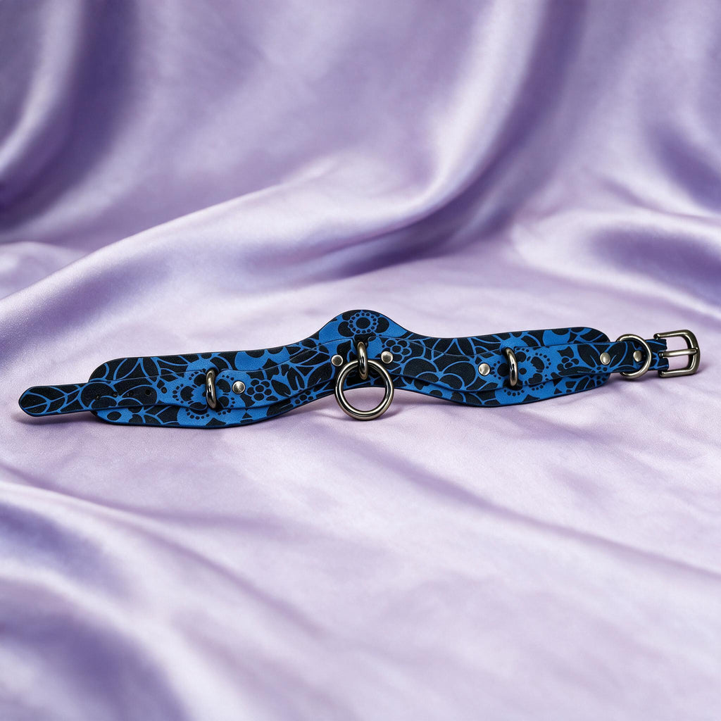 A blue and black patterned choker necklace with metal rings and a metal buckle is displayed on a purple fabric background.
