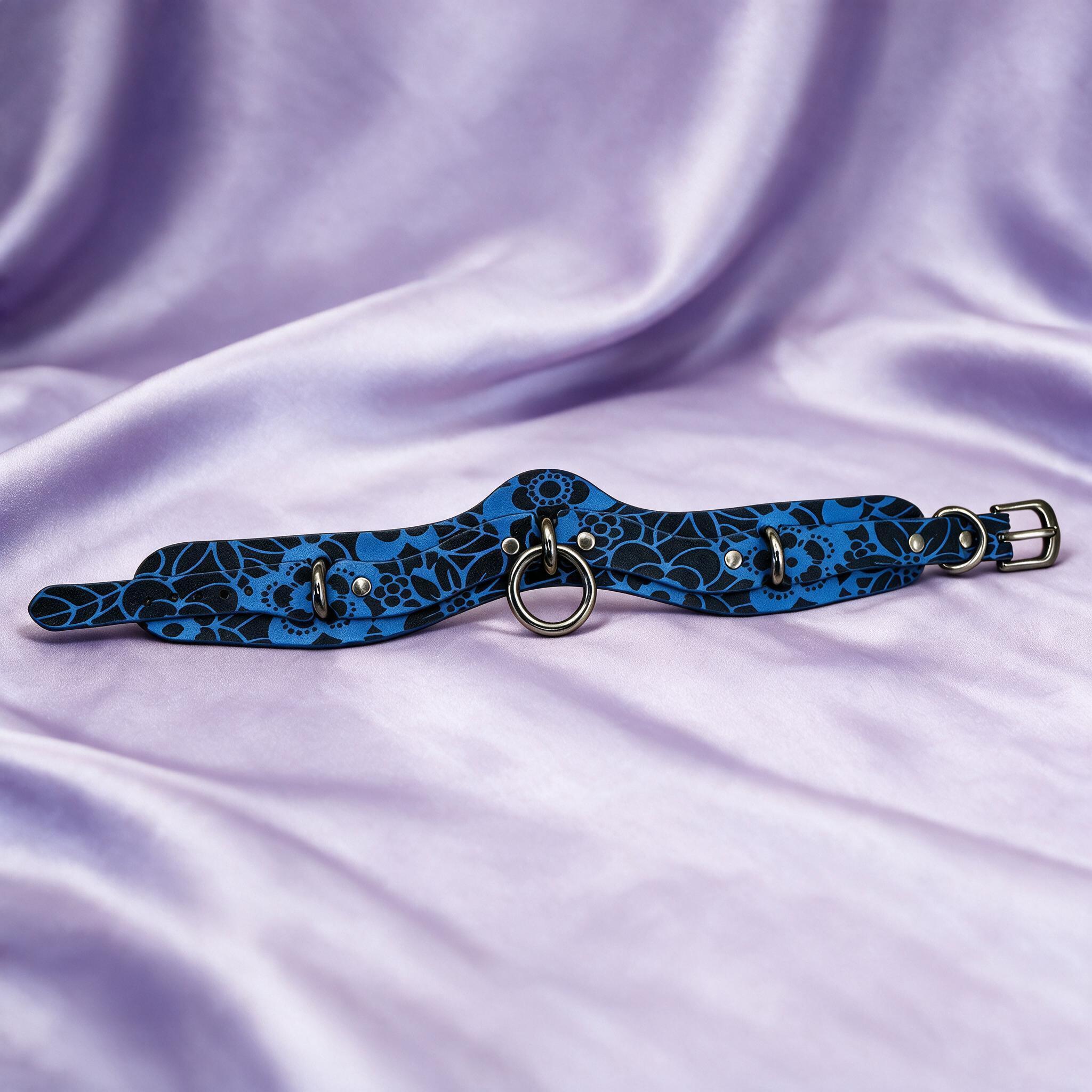A blue and black patterned choker necklace with metal rings and a metal buckle is displayed on a purple fabric background.