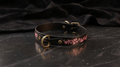 A black leather collar with a pink floral design on it, resting on a dark surface.