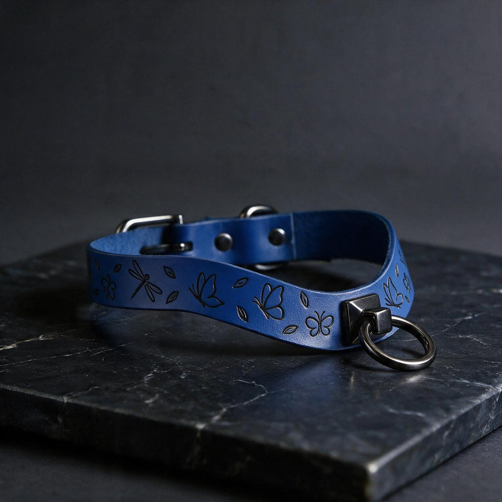 A blue leather collar with a metal ring and butterfly designs is displayed on a dark surface.