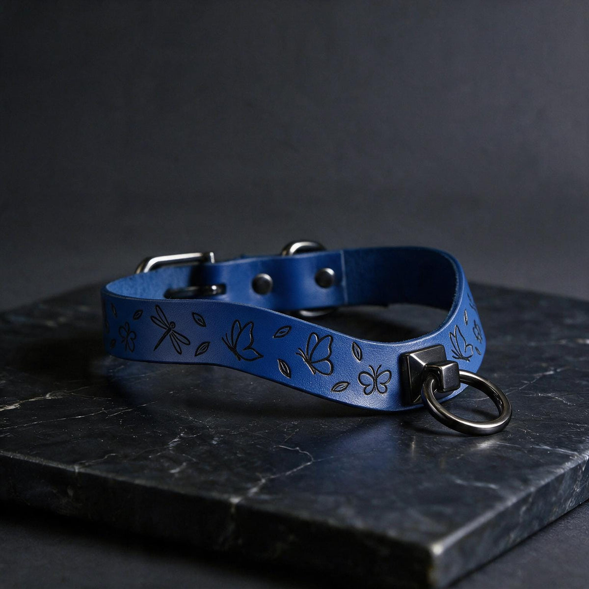 A blue leather collar with a metal ring and butterfly designs is displayed on a dark surface.