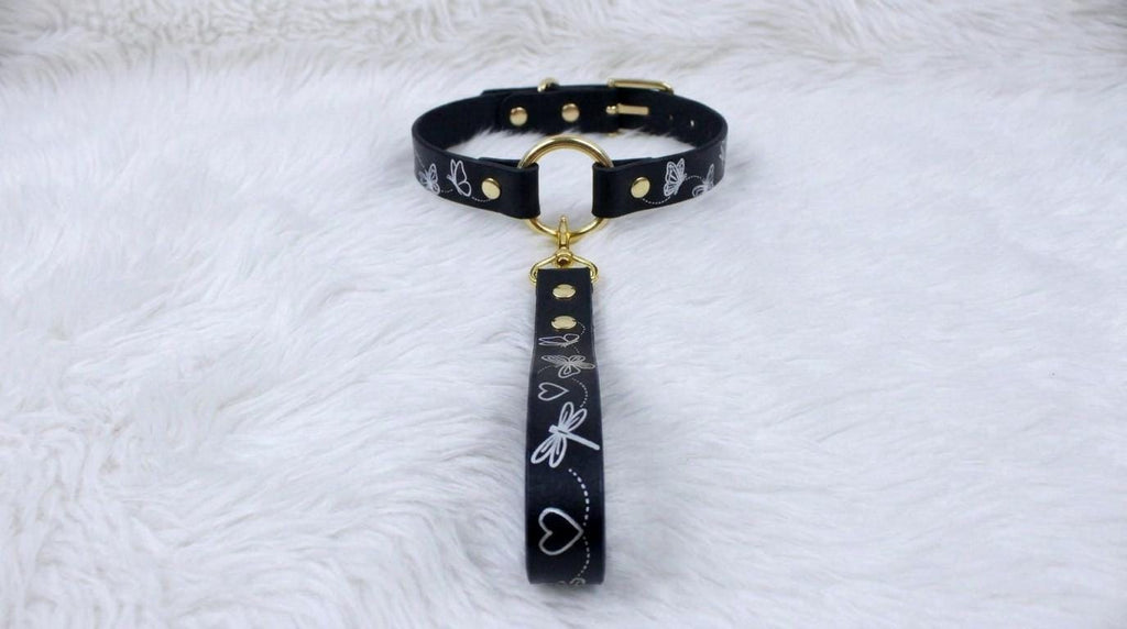 A black leather collar with a gold-colored buckle and a black handle with white designs is displayed on a white furry surface.