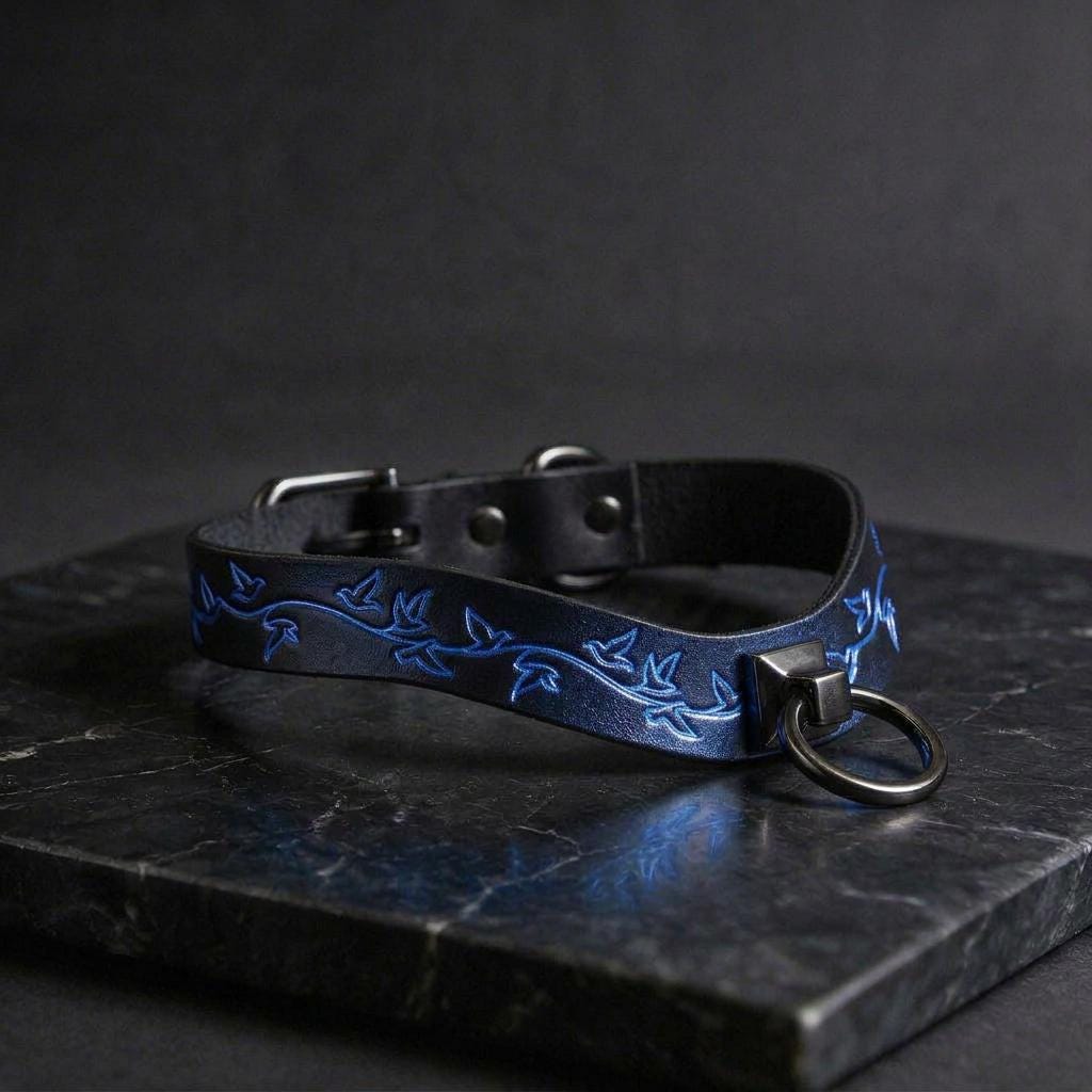 A black leather collar with a blue leaf design and a metal ring on the front, resting on a dark surface.