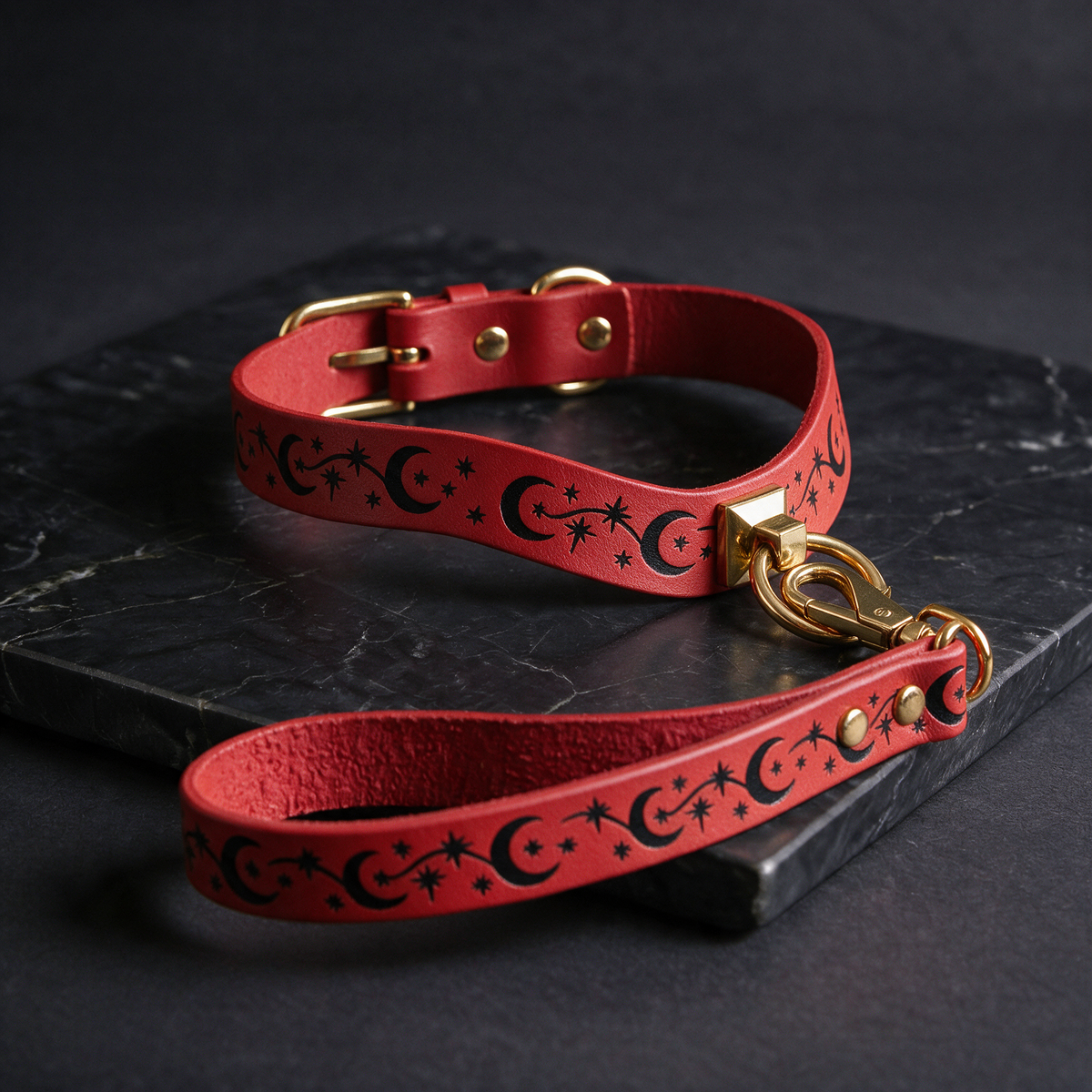 A red leather dog collar with a star and crescent design, and a matching red leash with a star and crescent design, both resting on a black marble surface.